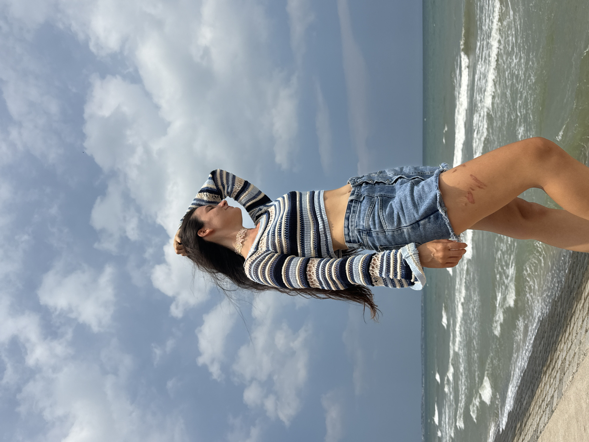 In an outdoor, medium-shot, full-body photograph, a young woman stands facing right, her left hand resting on the back of her head, right hand at her side. She wears a cropped, striped long-sleeved shirt and denim shorts. The setting appears to be a coastal area, with a concrete embankment in the foreground. The ocean stretches out behind her, with visible waves and a cloudy sky overhead. The specific location is Phu Thuy Ward, Vietnam. The woman's hair is dark and long, blowing slightly in the wind. Her eyes are closed, and her expression is relaxed. The overall scene is peaceful and serene.