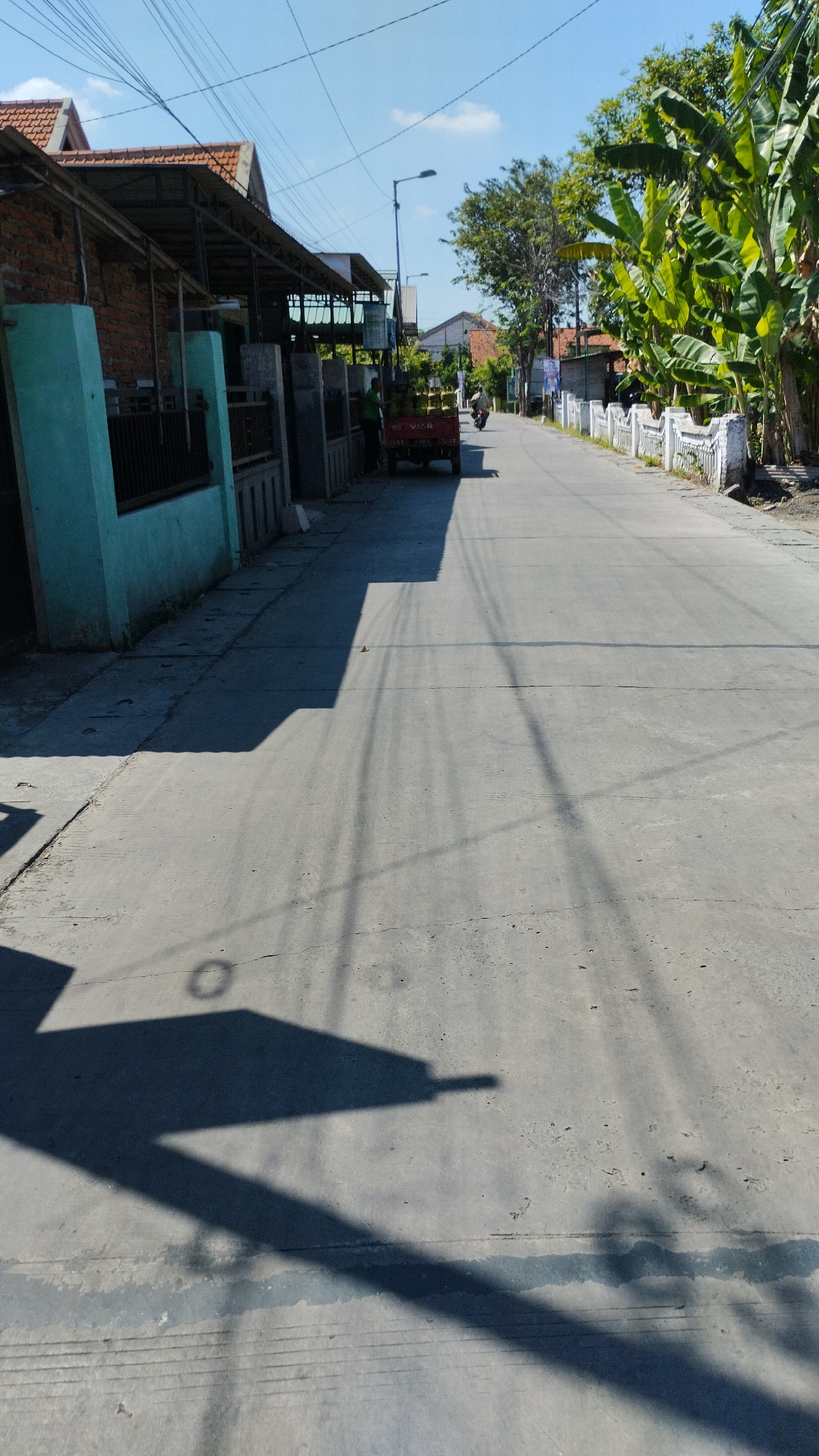 The image depicts a straight, paved road in a residential area of Sidodadi, Indonesia, under a clear blue sky with scattered clouds. On the left side of the road, there are houses with red tile roofs and brick walls, partially covered by awnings. A turquoise-painted wall is prominent in the foreground on the left. A red trailer, possibly attached to a vehicle not fully visible, is parked on the side of the road. A person in a green shirt is standing near the trailer. On the right side of the road, lush green banana trees line the street, and a white decorative fence runs along the property line. In the distance, more houses and trees are visible. The road surface shows tire marks and some cracking. Long shadows cast by overhead wires and objects on the left side of the road stretch across the asphalt. A motorcycle is seen in the middle distance on the right side of the road, with a rider. The overall impression is a sunny day in a tranquil Indonesian village.