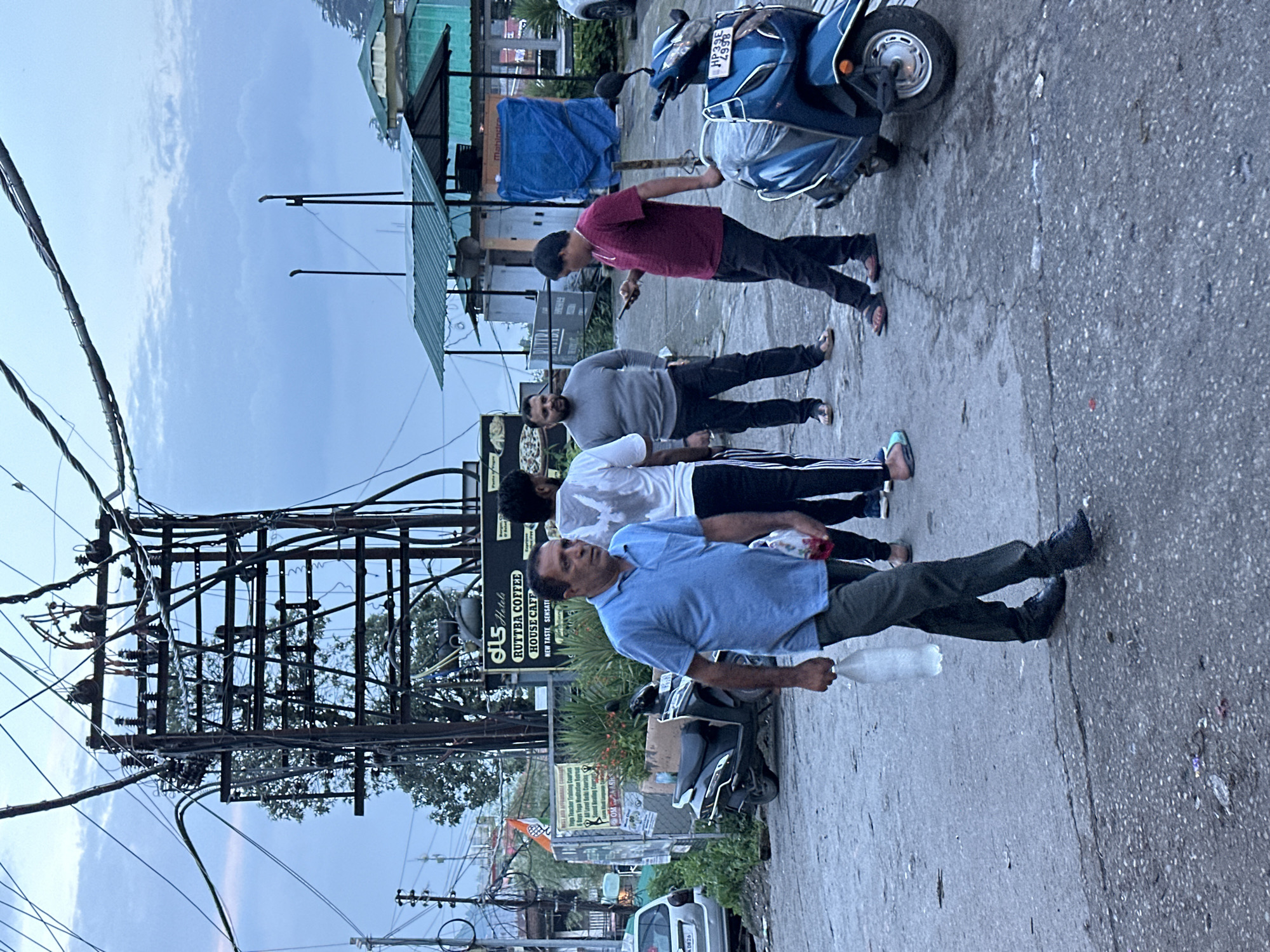 A medium-shot, eye-level, outdoor, full-color photograph captures a street scene at dusk. In the foreground, a man wearing a blue polo shirt and dark pants walks towards the left, carrying a clear plastic bag in his right hand. Behind him, to the right, a blue scooter is parked. Further back, a group of four men are visible. One man in a grey sweater and dark pants is looking down at his phone. To his left, another man in a white t-shirt and black and white striped pants stands. To the far left, a man in a maroon t-shirt and dark pants looks towards the right. The background reveals a complex network of overhead electrical wires and poles, some with transformers. In the distance, buildings with green roofs are present, and trees are visible. The sky is overcast with clouds. A scooter and a car are also parked on the side of the road. Signage for "DLS Hotels Ruttba Coffee House Cafe" is visible on a building.