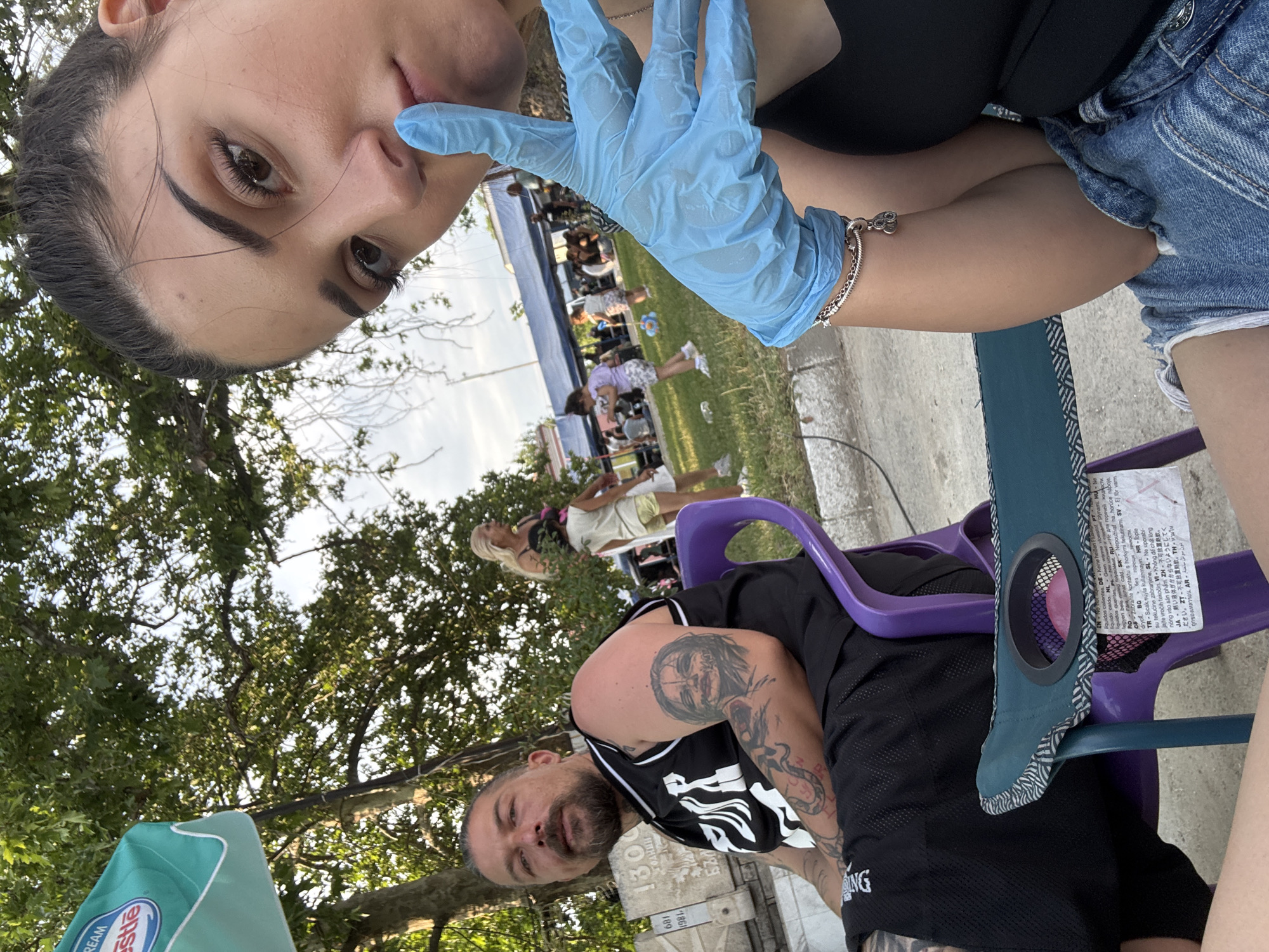 The image depicts an outdoor public gathering in Kostievo, Bulgaria. In the immediate foreground, a young adult female with dark hair, wearing a black tank top and denim shorts, is taking a selfie. Her right hand, covered by a light blue disposable glove, is positioned with the index finger to her lips in a "shhh" gesture. A silver charm bracelet, featuring what appears to be Olympic rings, is visible on her right wrist. She is seated in a low fabric camping chair with a built-in cup holder.
To her left, a middle-aged male with a beard and dark hair tied back is seated in a purple plastic chair. He wears a black sleeveless basketball-style jersey with white lettering and black shorts. His arms are heavily tattooed, with a distinctive detailed face on his right bicep and a serpentine or abstract design on his left forearm. He is looking towards the general direction of the camera. Behind him, a light-colored stone monument is visible, inscribed with Cyrillic text including "1300", "1981", and "БЪЛГА", consistent with a Bulgarian commemorative marker.
The background shows a public space with a mix of paved ground and grassy areas. An adult woman with blonde hair, dressed in a light-colored top and shorts, stands near a tree, looking to the right. Several children are present, including one in a white top and yellow shorts, playing on the grass. Further in the background, under multiple blue tarpaulin tents, a crowd of people is gathered, indicating a community event or market. The scene is illuminated by natural daylight, suggesting late afternoon or early evening. A green and white umbrella, partially visible in the upper left, displays "NESTLE" and "DREAM" branding.