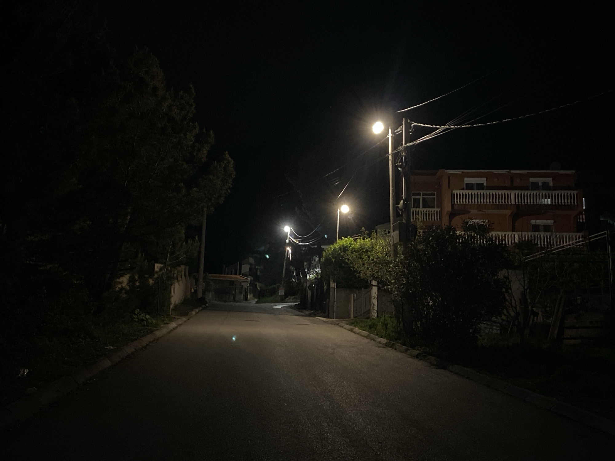 The photograph depicts a street in Šušanj, Montenegro, at night. The scene is illuminated by streetlights, casting a glow on the asphalt road. On either side of the road are buildings, trees, and vegetation. The building to the right is two stories tall with a balcony. Power lines can be seen overhead. The image is taken from a low vantage point, looking down the road.