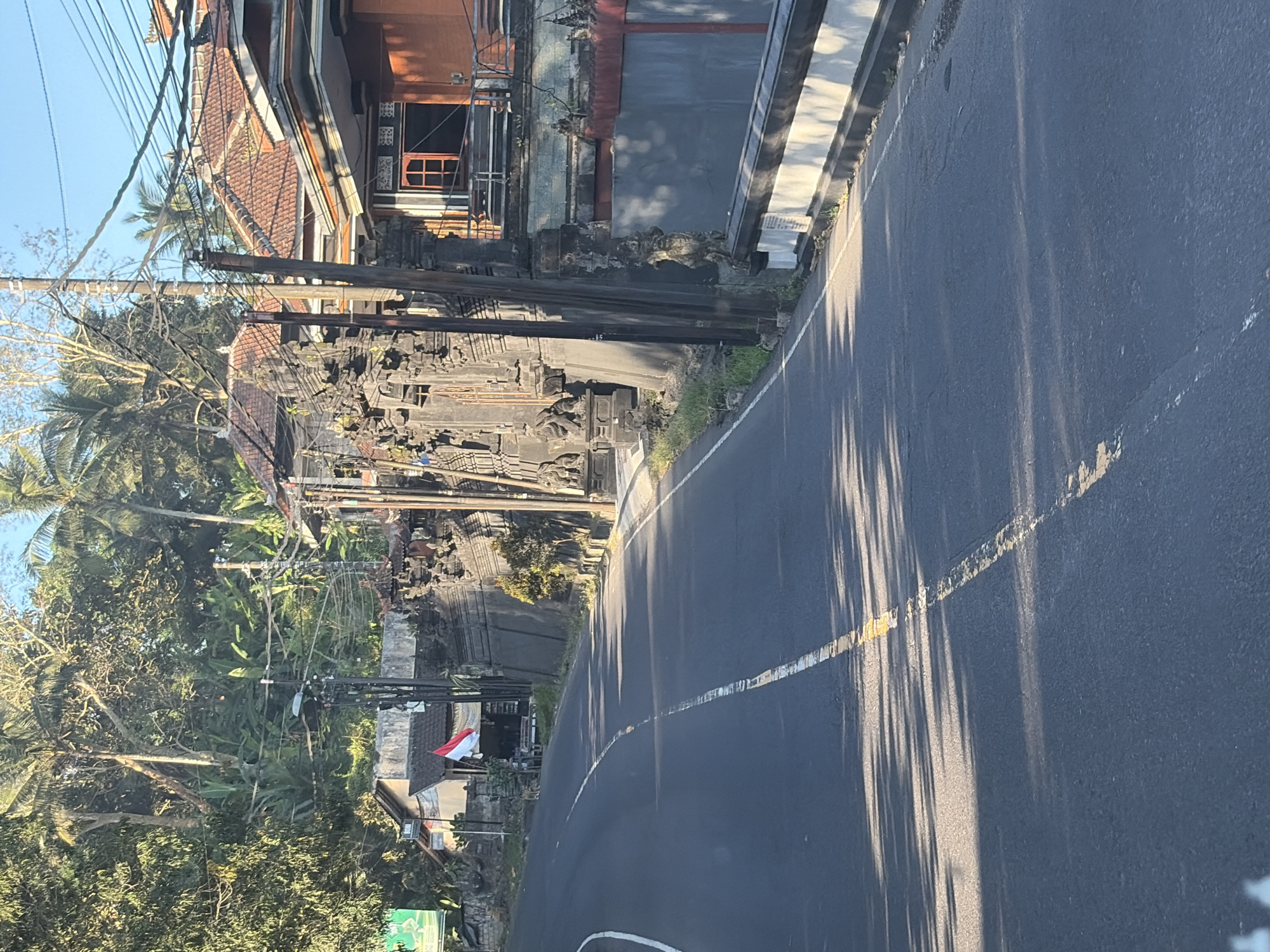 This is a photograph of a paved road curving to the left, bordered by vegetation and buildings. A white line is painted down the center of the road, and a yellow line runs along the right edge of the lane. In the background, to the right of the road, there is a stone wall with intricate carvings. Behind the wall, a building with an orange facade and tiled roof is visible. To the left of the road, there are more stone structures and some buildings with red-tiled roofs. An Indonesian flag is displayed on a flagpole in front of one of these buildings. Power lines crisscross the scene, supported by wooden poles. Lush green trees and palm trees dominate the background, suggesting a tropical environment. The lighting indicates it is daytime, with shadows cast by the trees on the road. The scene appears to be a street in a residential or commercial area, possibly in Karangasem, Indonesia, given the architectural style and flag.
