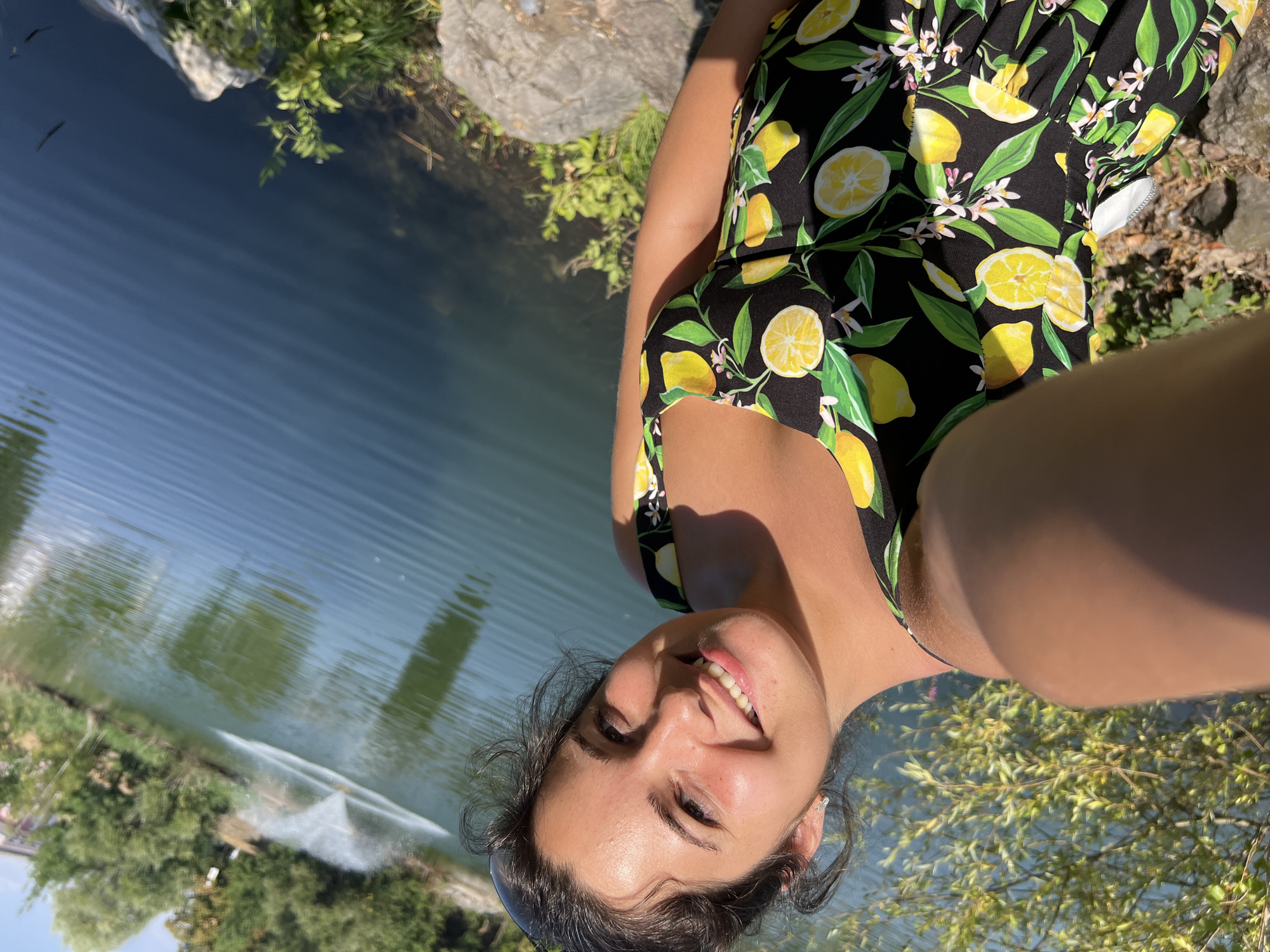 This is a vertical, low-angle, outdoor selfie taken in Sofia, Bulgaria. The main subject is a young woman with dark hair and a bright smile, wearing a black swimsuit with a lemon pattern. She is positioned in the foreground, with her face tilted upwards towards the camera. The background features a body of water, likely a pond or lake, with some greenery and rocks along the edge. A fountain is visible in the water in the distance, creating a spray. The scene is illuminated by bright sunlight, suggesting a clear day. The overall mood is cheerful and relaxed, with the woman's smile conveying happiness. The time of day appears to be daytime, based on the lighting conditions. There is no visible text in the image.