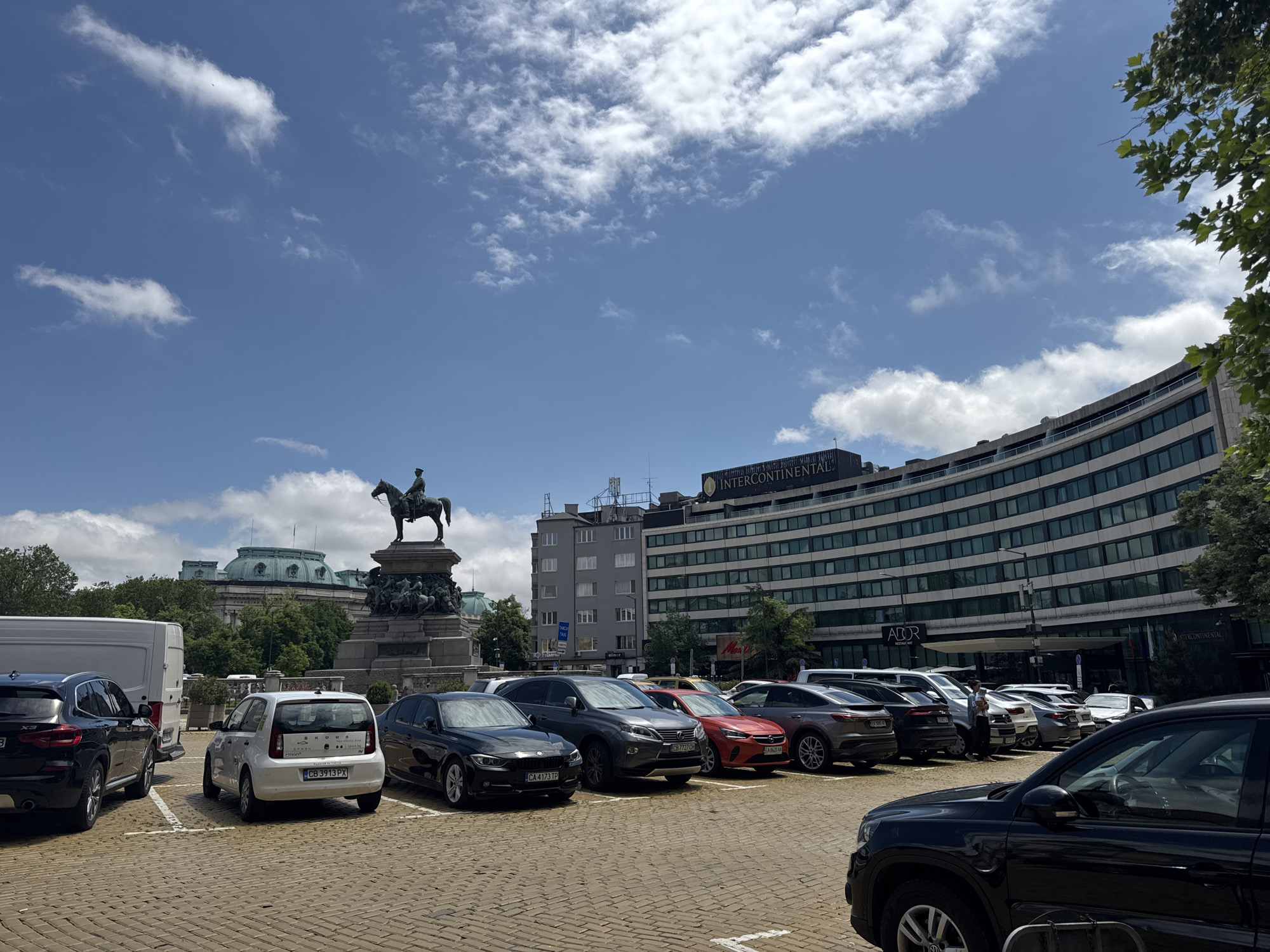 The image presents a bright, sunny daytime scene in Sofia, Bulgaria, showcasing a mix of historical landmarks and modern city elements. The sky is a clear blue, adorned with scattered white cumulus clouds.
In the foreground, a paved parking lot, distinguished by its light-brown, irregular stone surface, is densely packed with a variety of parked cars. These include several black SUVs and sedans, a white Skoda hatchback (license plate "CB 3913 PX") with "SKODA" and "sparx.bg" visible on its rear window, a white van, a grey SUV (license plate "CB 7721 CH"), and a red car (license plate "CA 8261 KH"). Another black sedan with the license plate "CA 4173 TP" is also visible. One person is seen standing near the white van in the mid-right.
The midground features prominent historical structures. On the left, a large equestrian statue, likely the Monument to Tsar Liberator, depicts a man on horseback atop a grand, ornate stone pedestal. Behind the statue and partially obscured by green trees, a classical building with a distinctive green-domed roof can be seen, which appears to be the National Assembly building. Further right, other multi-story urban buildings are visible.
The background is dominated by a large, curved, multi-story building, identified by the prominent "INTERCONTINENTAL" sign at its top and "AD OR" written below it. This building has many windows and a modern aesthetic. Green foliage from trees frames the upper right and left edges of the photograph. The overall scene depicts a lively urban environment where historical monuments and contemporary city life coexist under a clear summer sky.