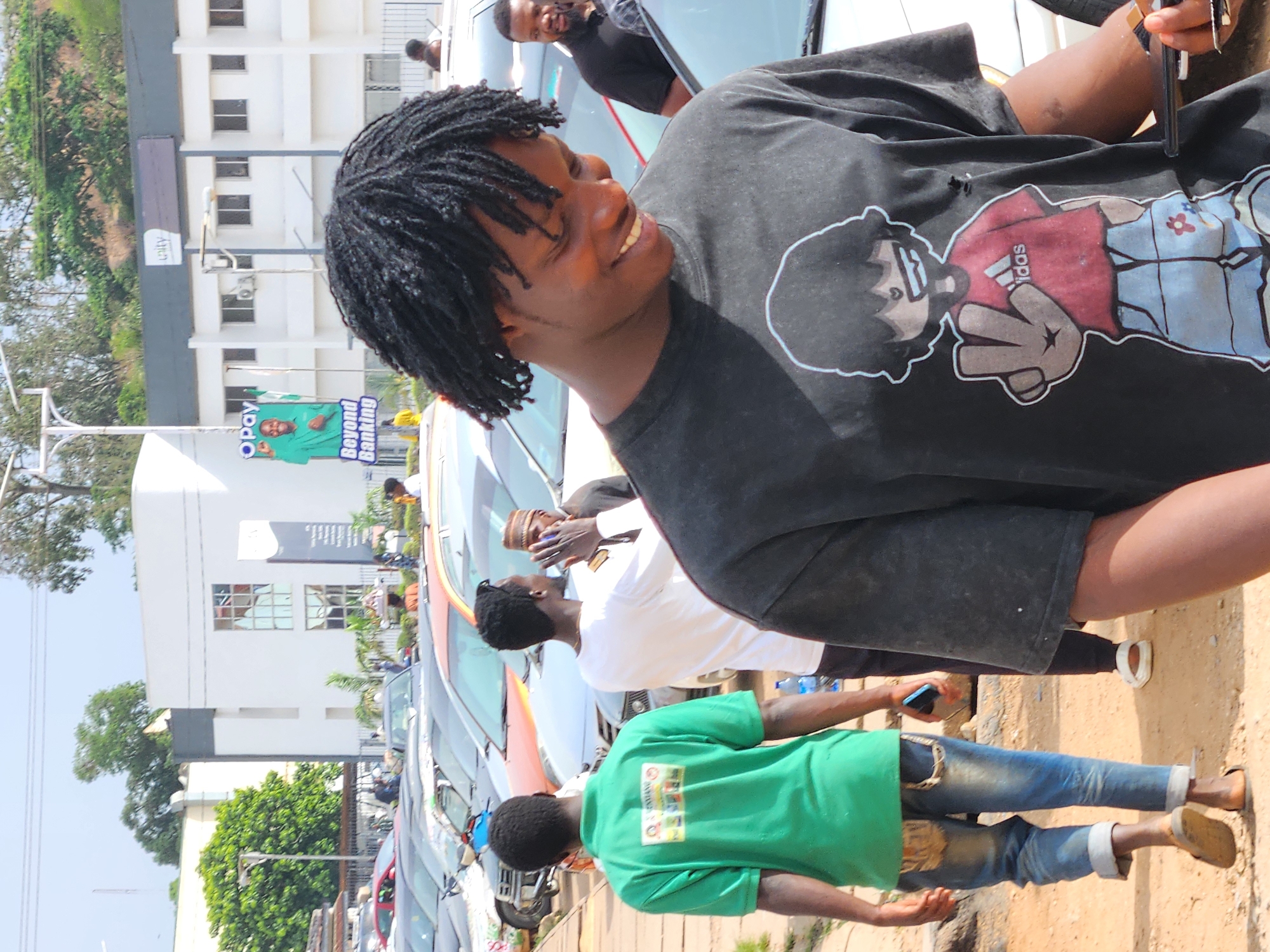 The image shows a street scene. In the foreground, a person with dreadlocks is wearing a black t-shirt with a cartoon graphic. They are smiling and holding an object in their right hand. Behind them, other people are visible, including one in a green t-shirt and another in a white t-shirt. Cars are parked along the street. In the background, there are buildings, trees, and a sign that reads "Opay Beyond Banking." The image is taken during daylight. The location is Unknown, Unknown.