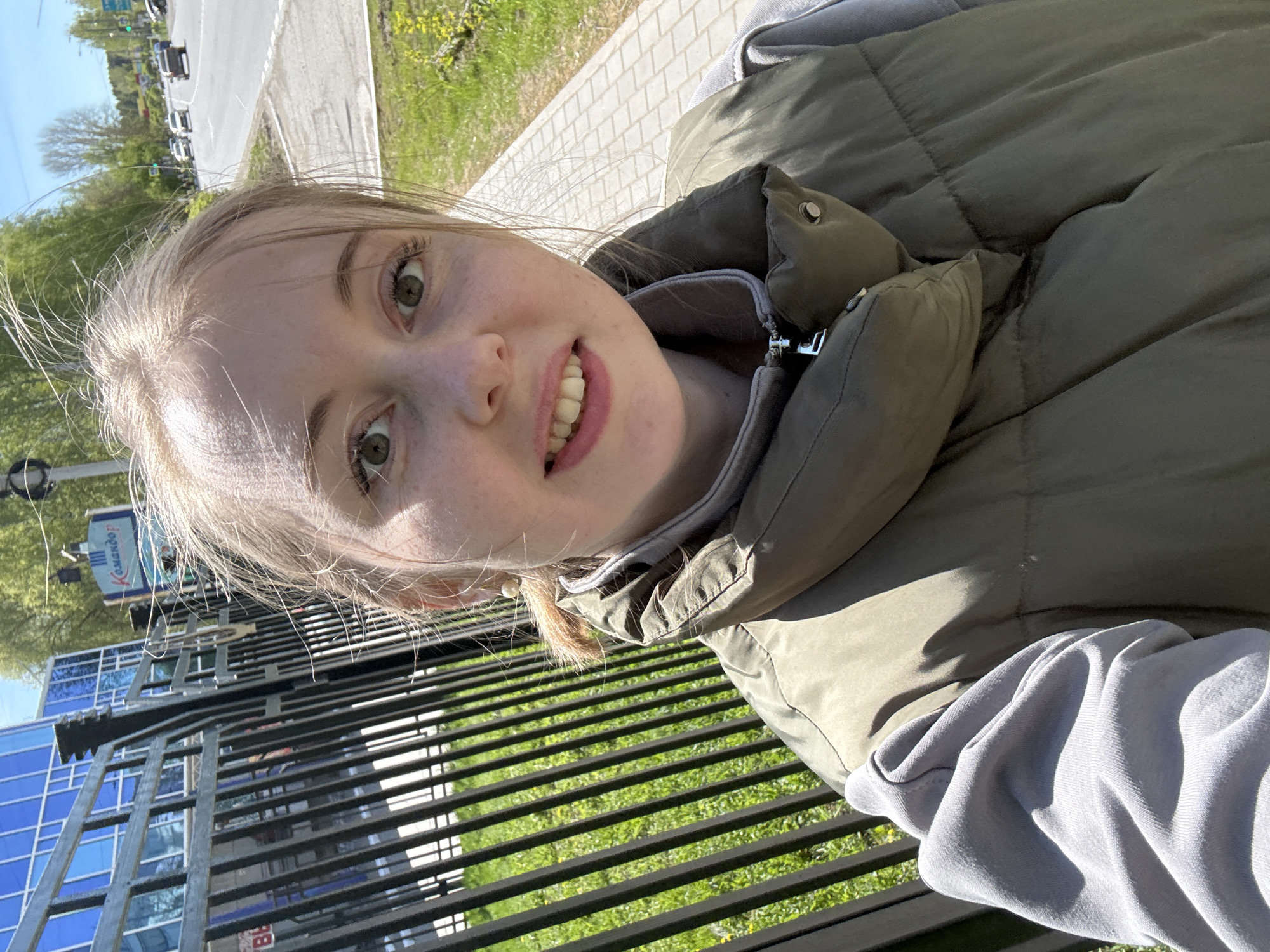 A young woman takes a selfie outdoors. She is wearing a khaki-colored puffy vest over a light gray sweatshirt. She is standing behind a black metal railing, with grass visible on the other side. Beyond the railing is a modern blue and glass building, with a sign in Russian visible on top. A road and some trees are visible in the background. The woman is smiling slightly at the camera. The location appears to be in the city of Krasnodar, based on the sign visible in the background.