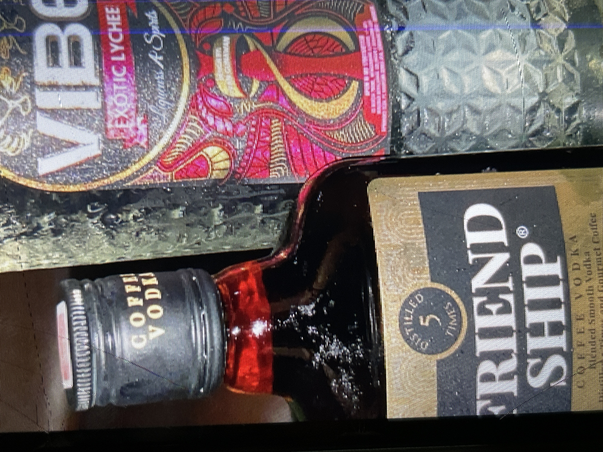 The image shows two bottles of alcohol. The bottle in the foreground is a dark brown glass bottle with a black cap. The cap has text that appears to read "COFFI VODKA". The bottle has a label that says "FRIEND SHIP" in large white letters, with "DISTILLED 5 TIMES" above it. The bottle also states "COFFEE VODKA" and "Blended Smooth Vodka". The bottle on the right is a clear glass bottle with a label that has a pink and red design. The text on this label reads "VIBE" in large white letters and "EXOTIC LYCHEE". There is no discernible city, location, or time context within the image itself.