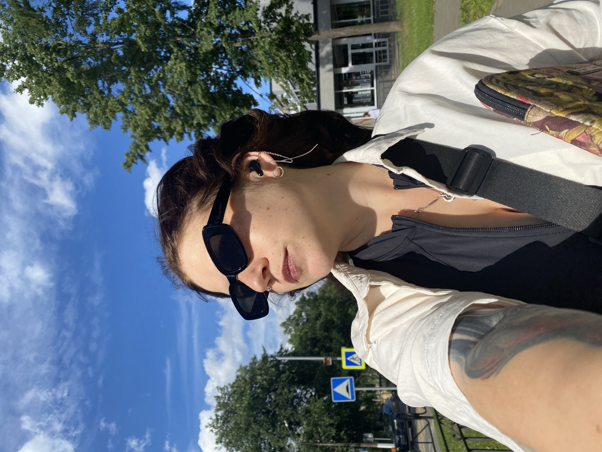 A medium shot taken outdoors on a sunny day shows a young woman from the chest up, looking to her right. She has dark hair pulled back, is wearing black sunglasses, and has a nose ring and a delicate chain earring with a long string. She is wearing a black zip-up top and an unbuttoned white collared shirt over it. A black strap goes across her chest, and a patterned bag with a zipper is visible on her right side. Her left arm is partially visible, showing a large tattoo. In the background, there are trees, a building, a sidewalk, a street, and traffic signs. The scene appears to be located in the city of Chaikovsky, Russia, indicated by the presence of traffic signs commonly found in Russia.