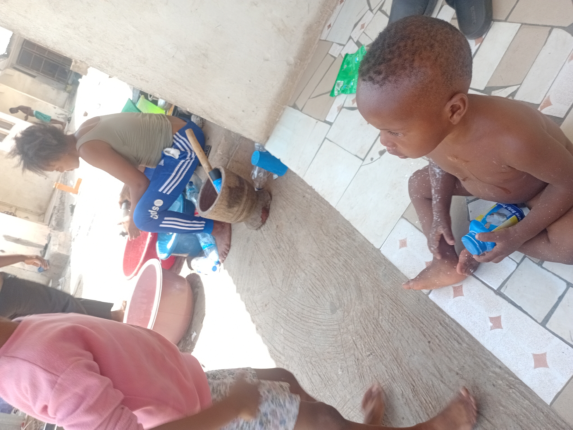 The image captures an outdoor scene in bright daylight, likely a courtyard or open area adjacent to a building in Lokoja, Nigeria.
In the foreground and central focus, a young child, possibly a boy, is seen bare-chested and squatting on patterned tiles. His skin is wet, and he holds a blue plastic bottle, partially legible as "Frisky Baby Powder," suggesting he is either bathing or playing with water.
To the left, a woman or teenager with dark, short hair is bending forward. She wears a greenish-brown sleeveless top and blue athletic pants with white stripes. Near her are a traditional wooden mortar and a pestle (or similar wooden tool), along with another blue plastic bottle and a white striped towel.
Another person, wearing a pink long-sleeved top and patterned shorts/skirt, is partially visible in the bottom left, with their bare feet also in the frame. In the background, a person's arm and leg are visible, and a building with a window can be seen.
Other objects scattered on the ground include red and brown plastic basins and a green plastic sheet/bag. The ground is a mix of white tiles with small brown/orange diamond patterns and a rough concrete slab. Black sandals are visible in the top right corner. The scene depicts a casual, everyday activity, possibly involving household chores or children's play.