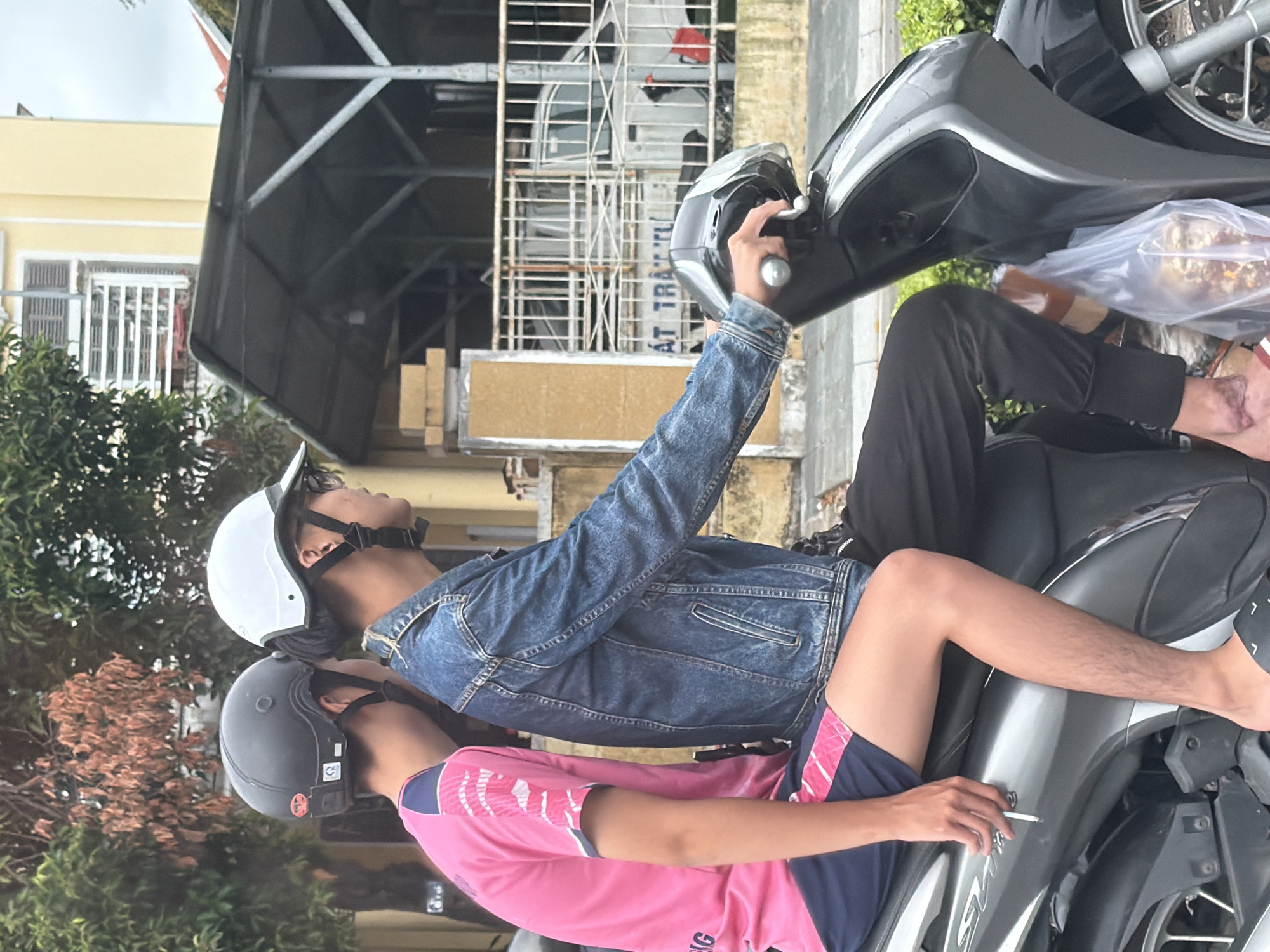 Two individuals are seated on a grey and black scooter, traveling on a street in Phú Xuân Ward, Vietnam. The person in the rear is wearing a pink t-shirt with blue and white trim and blue shorts with pink accents. The person in the front is wearing a denim jacket, black pants, and a white helmet. The driver is wearing glasses and is holding the scooter's handlebars. The background shows trees, a yellow building, a corrugated metal roof structure, and a white metal fence with a white car behind it. The ground appears to be asphalt. The scooter has visible wheels and bodywork.
