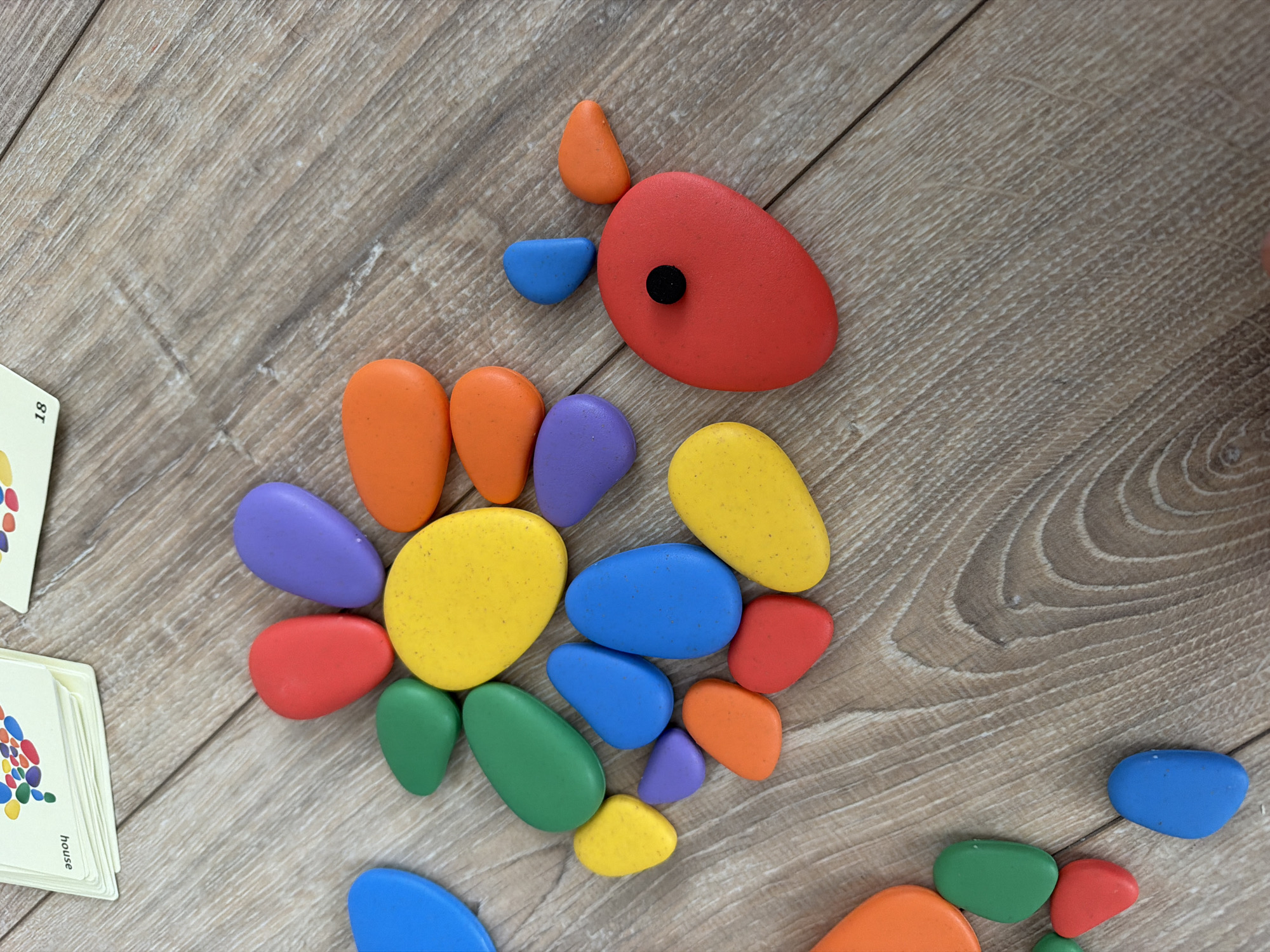 The image shows a collection of colorful, pebble-shaped toys scattered on a wooden floor. The toys are predominantly red, orange, yellow, green, blue, and purple. Some of the toys are arranged in a circular pattern resembling a flower. There are also two cards visible in the upper left corner of the image, one with the word "house" and another with the number "18." A red pebble toy with a black dot in the center, resembling an eye, is also present, along with two smaller blue and orange pebbles positioned to look like ears or fins. The lighting suggests an indoor setting, likely a child's play area. The image does not provide any information about the city of Chaikovsky, Russia, or any specific location within it.