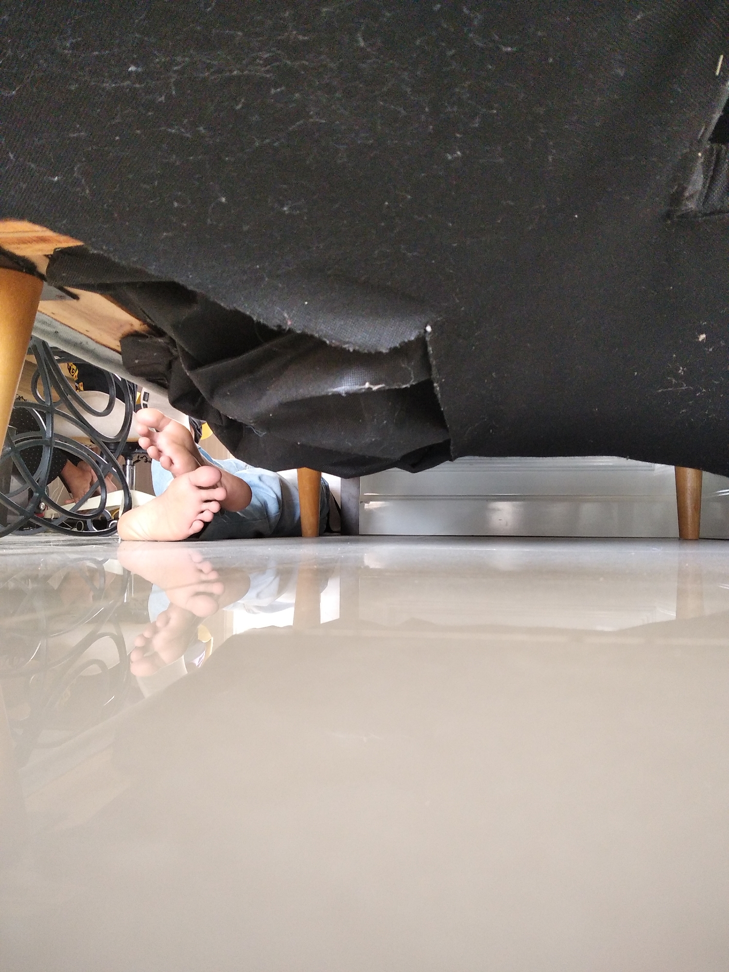 The image depicts a low-angle view from beneath a piece of furniture, possibly a sofa or bed, showing the feet of a person lying on a polished floor. The person is wearing light blue pants. The furniture's dark fabric underside is visible, along with wooden legs. In the background, electrical cords and a metal structure, possibly a chair, are also visible. The floor is highly reflective, showing a clear mirror image of the feet and the surrounding area. The scene appears to be indoors. The specific location in Kaliwates, Indonesia is not discernible from the image.