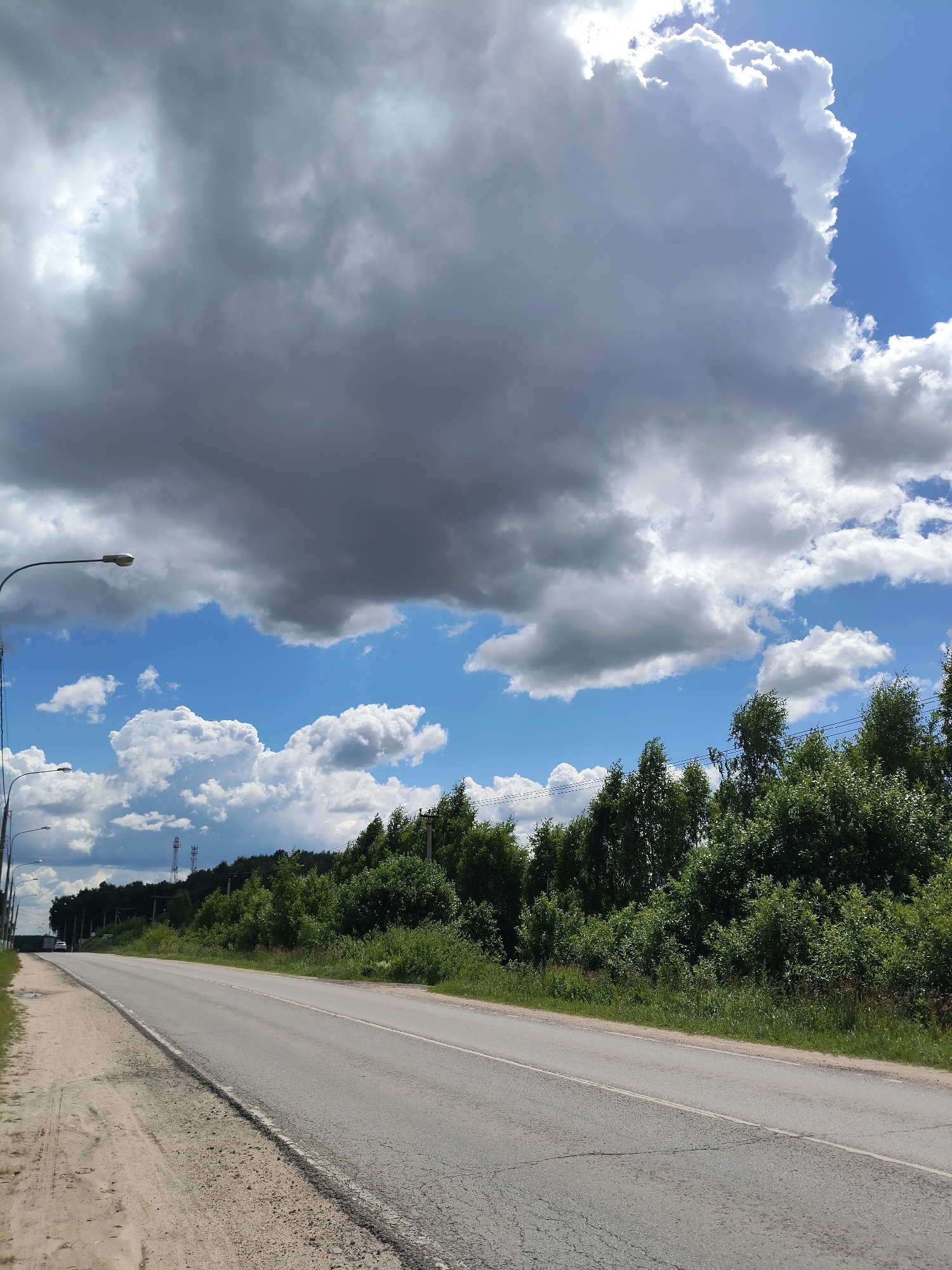 The image shows a road curving into the distance, bordered by a sandy verge on the left and a dense line of green trees on the right. The sky is a mix of bright blue with scattered white clouds and large, dark grey storm clouds. There are streetlights visible along the left side of the road. In the distance, on the right side of the road, there are telecommunication towers visible above the treeline. The road surface is asphalt and appears to be in fair condition with some cracks. A white car is visible in the distance on the road. The overall impression is of a summer day with developing weather conditions. The time of day appears to be daytime, possibly late morning or afternoon, given the brightness of the sky. The location is identified as Khodaeovo, Russia. There is no visible text in the image.