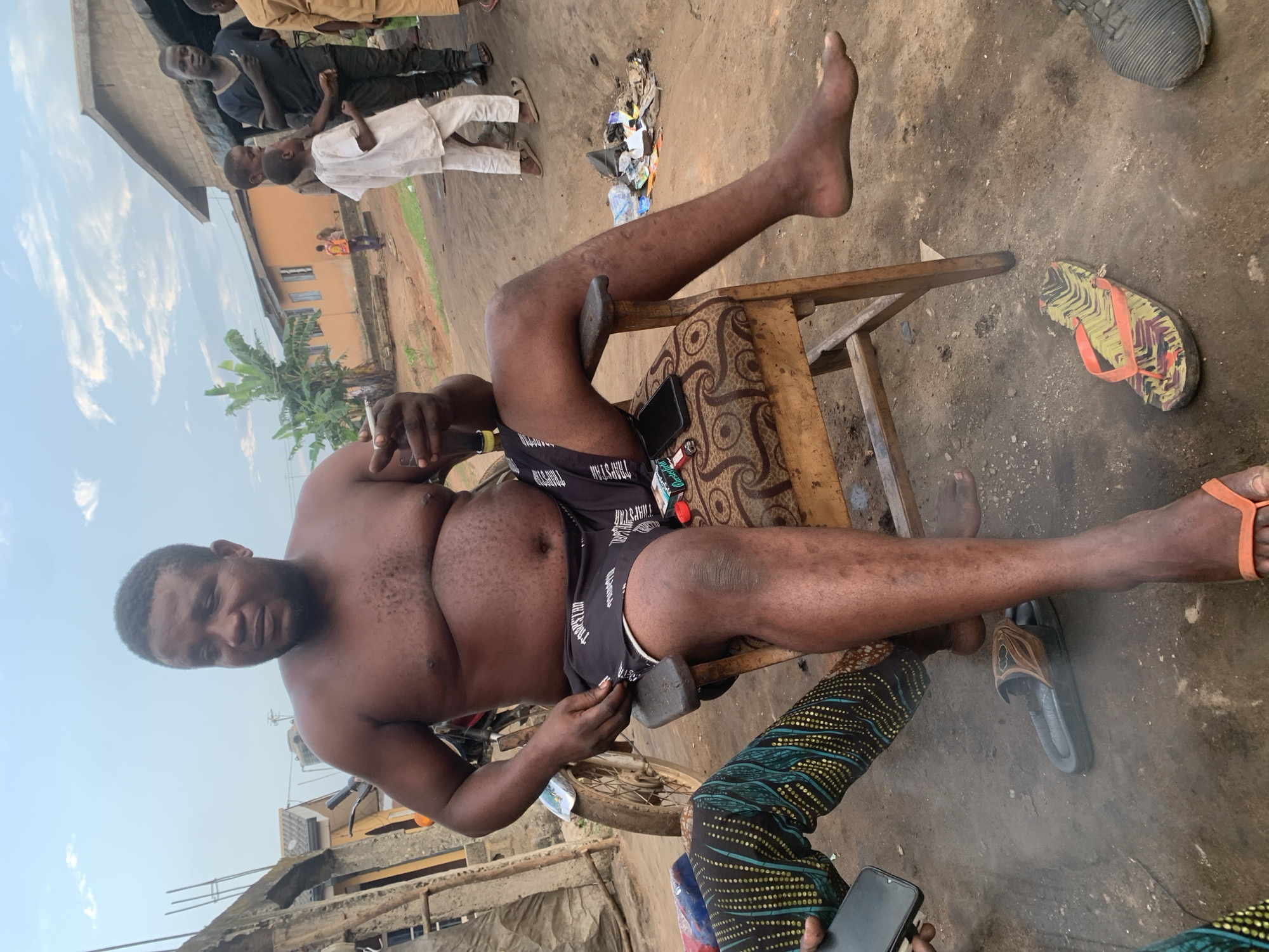 In the city of Shagamu, Nigeria, a shirtless man with a dark complexion is seated on a wooden chair with a patterned cushion. He is wearing black shorts with a white pattern and appears to be holding a cigarette and a bottle. To his left, another person's legs are visible, wearing colorful patterned leggings. In the background, children and adults are present, with buildings and a motorcycle visible. The ground is made of dirt and debris. A pair of flip-flops lies on the ground near the seated man's feet.
