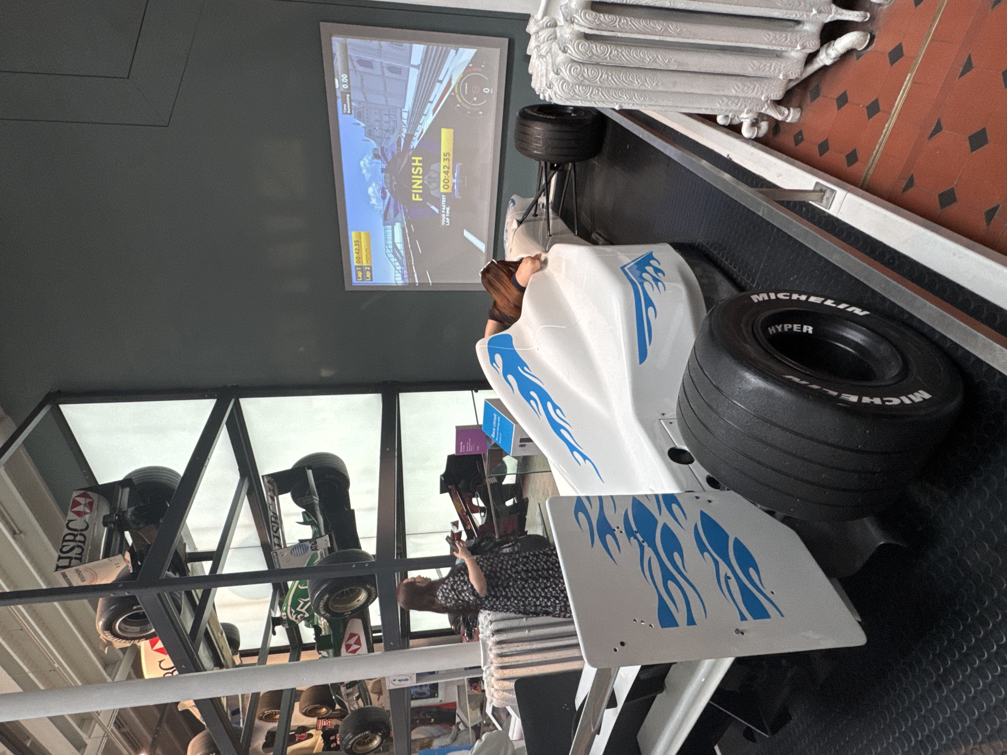 A woman, with long brown hair and wearing a black and white floral dress, stands to the left of a white and blue racing simulator in the City of Edinburgh, United Kingdom. The simulator, designed to resemble a Formula 1 car, is positioned on a black mat. Behind the simulator, a large screen displays a racing game, with the word "FINISH" prominently visible. To the left of the woman, several racing cars are displayed on tiered shelving units, some featuring sponsor logos such as HSBC and Bridgestone. A white, decorative radiator is located to the right of the simulator. The floor is a mosaic of red and black tiles.