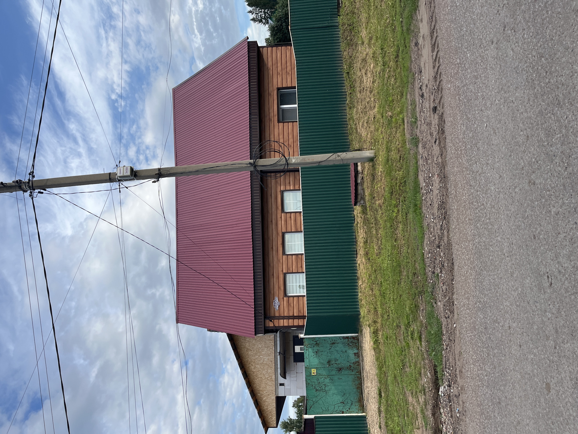 The image depicts a residential street scene in the city of Ufa, Russia, with a focus on a wooden house and its surroundings. The house, constructed of horizontally laid logs, features a prominent, dark red corrugated metal roof. A dark green metal fence runs along the front of the property, with a section of a lighter, aged green metal gate visible on the left. Behind the fence, a patch of green grass and dirt is present. A concrete utility pole stands in front of the house, to the right of center, with electrical wires strung across the frame and a small white meter box attached to the pole. Part of another building, with a lighter colored roof and white block construction, is visible to the left of the main house. The sky above is partly cloudy with white clouds against a blue sky. The foreground shows a paved road.