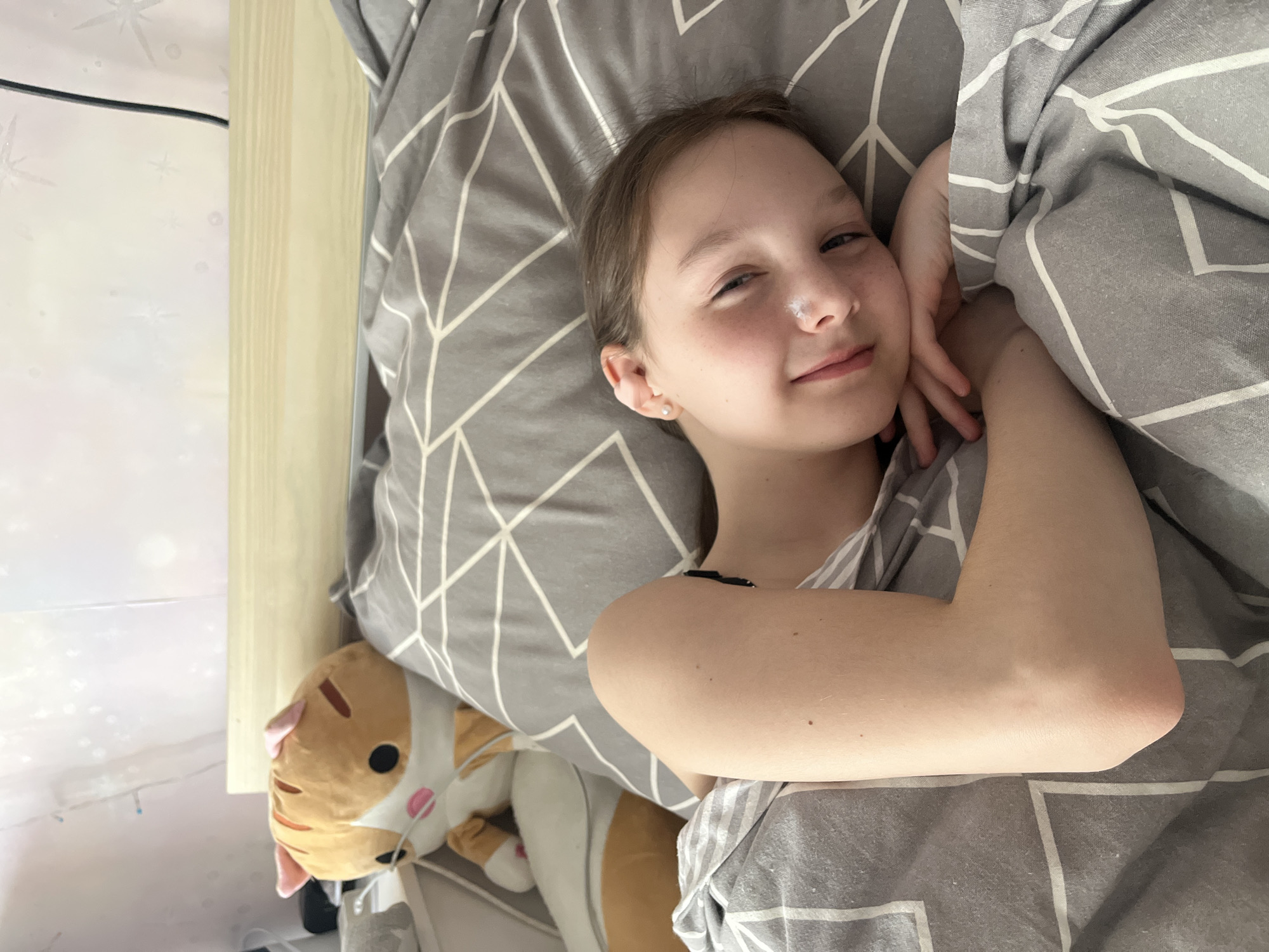 The image, rotated 90 degrees clockwise, captures a young girl lying in bed. She appears to be in her early teens, with light brown hair, a fair complexion, and visible freckles. A small, light-colored smudge or spot is on her nose. She is wearing a light-colored, possibly striped, top with thin straps and a small stud earring in her left ear. Her expression is relaxed and playful, with a subtle smile and her left eye partly closed, as she looks directly at the camera. Her left hand is gently resting near her face.
She is partially covered by a grey duvet with a distinct white geometric pattern of interlocking lines, and her head rests on a matching pillow. To the lower left of the frame, a large, orange and white striped cat plush toy with pink ears and nose is visible, nestled close to the bed. In the background, behind the girl, a light-colored wooden surface (likely a headboard or bedside table) is present, along with a light wall adorned with faint, star-like patterns or decorations. A thin black electrical cord runs along the edge of this background.
The setting is an indoor bedroom, suggesting a moment of rest or waking up. The soft, natural lighting implies it is daytime, possibly morning. No specific visual cues in the image indicate the provided location of Chaikovsky, Russia. There is no visible text in the media file.