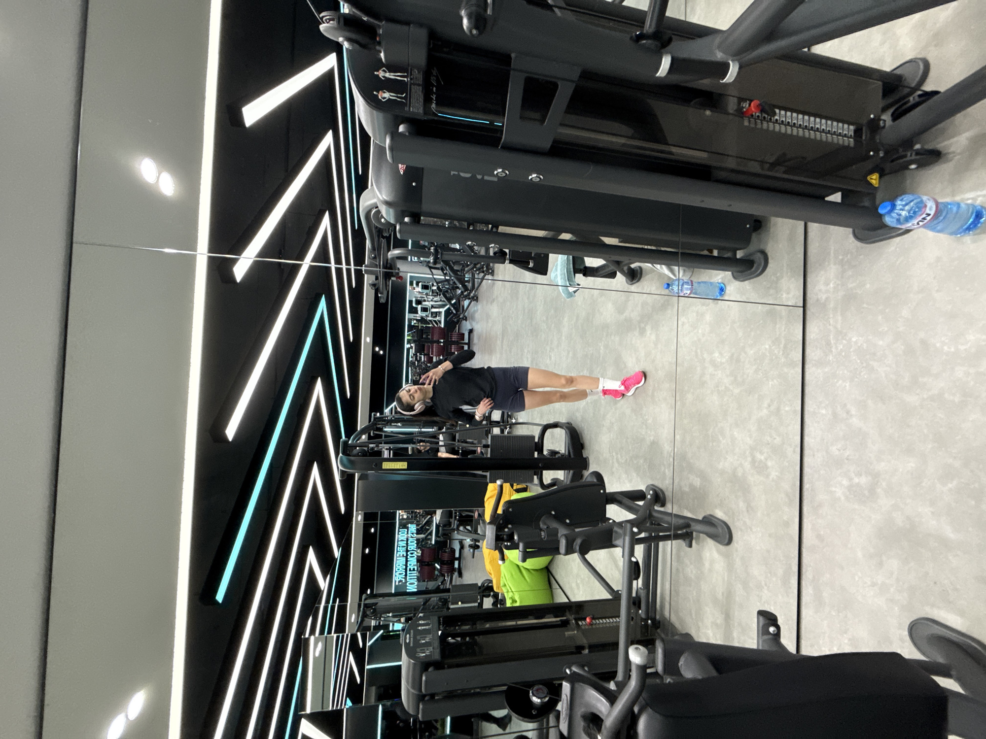 The image depicts the interior of a gym, likely in Sofia, Bulgaria. A woman in athletic attire (black top, dark shorts, pink sneakers) is visible lying on a bench, seemingly using her phone or listening to music with headphones. Gym equipment, including weight machines and cable stations, fills the foreground and background, their reflection visible in the large mirror on one side. The interior design features a modern aesthetic with geometric patterns of white and blue neon lights. A water bottle sits on the floor near one of the machines.