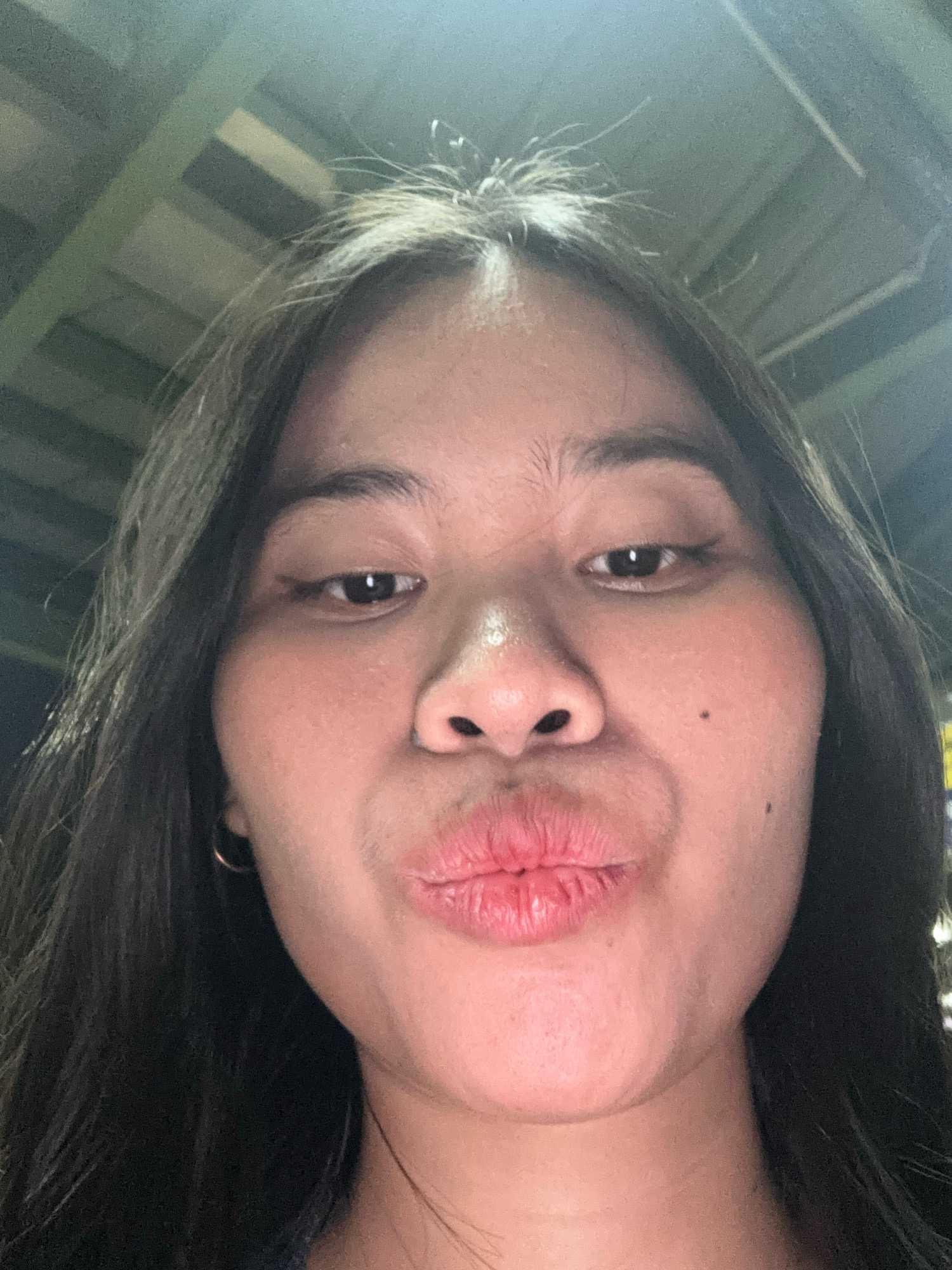 A low-angle, close-up selfie shows a young woman's face from the chin up, with her lips pursed in a kissing gesture. She has long, dark hair framing her face. Her skin is light brown, and she has a small mole on her right cheek. A thin hoop earring is visible on her left ear. The background is out of focus, suggesting an indoor setting with artificial lighting, possibly a ceiling with metal beams. The image was taken in East Jakarta, Indonesia.