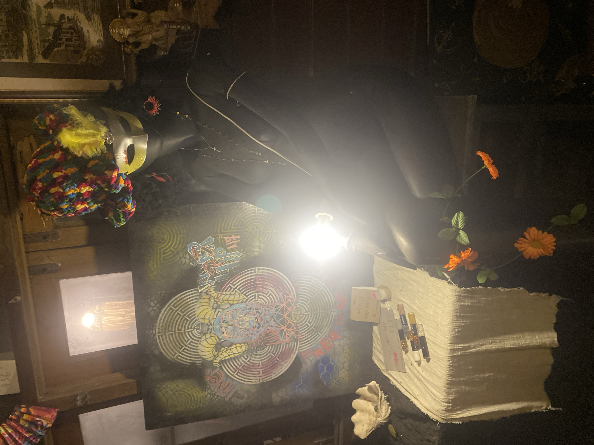The image depicts an interior scene in what appears to be a shop or studio in Baan Don Sai, Thailand. The lighting is dim, and the view is from an elevated angle looking down. The most prominent elements include: a mannequin head wearing a colorful hat and mask; a large, dark, draped object resembling clothing or fabric; a canvas with intricate, colorful geometric designs; a stack of what appear to be paper or textile materials; and a few artificial orange flowers. There are also various smaller objects and art pieces visible in the periphery. The location is identified by a small note that reads "THE HANG @ TREE HOUSE" indicating that is likely the name of the location. The overall impression is one of a cluttered but creative space, possibly associated with art, fashion, or craftsmanship.