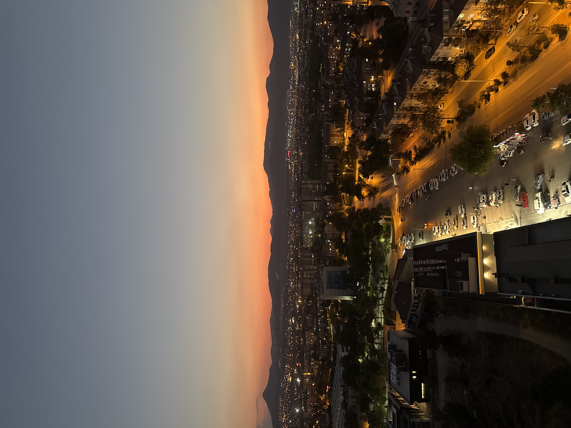 The image captures a wide-angle view of a city at dusk, with a vibrant orange and purple gradient sky transitioning to a deep blue. In the foreground, the rooftops of buildings and a parking lot with numerous cars are visible, illuminated by streetlights and building lights. A main road runs through the scene, with cars driving and parked along its sides.
In the middle ground, the city stretches out with a dense collection of buildings, many of which are lit up, indicating evening activity. Trees and green spaces are interspersed throughout the urban landscape. A prominent building with a large sign is visible in the mid-left. Further in the background, a mountain range forms the horizon, silhouetted against the colorful sky. The overall impression is of a bustling urban environment settling in for the night.
The scene is consistent with an aerial view of the city of Konya, Turkey, during twilight. Specifically, the layout and building architecture are indicative of urban areas within Konya. The presence of a major road, residential and commercial buildings, and the surrounding landscape align with typical cityscapes in this region of Turkey. While no specific landmarks are definitively identifiable without further context, the general urban characteristics strongly suggest Konya.