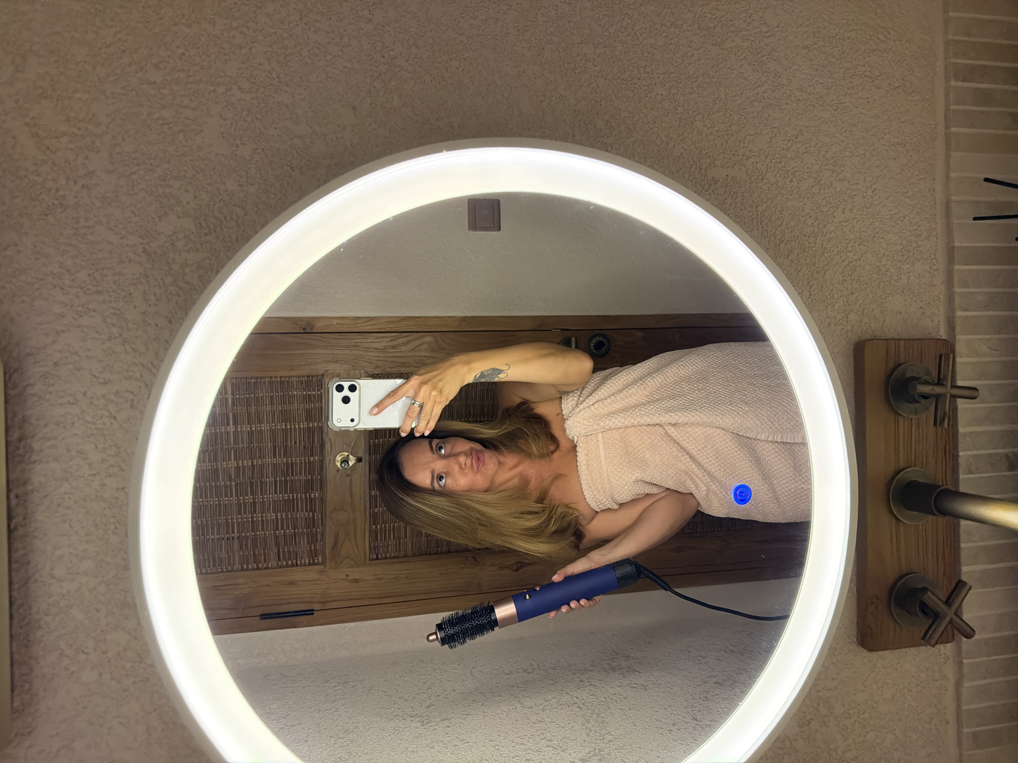 A woman is taking a selfie in a large, illuminated circular mirror. She is wrapped in a light pink towel and holding a blue and black hair styling tool. She is looking at her phone, which is also visible in the mirror. The background appears to be a bathroom with wooden accents and textured walls. A small blue light is visible on her towel in the reflection. The image appears to be taken indoors, and there are no indications of time of day or weather. The overall mood is casual and relaxed.
