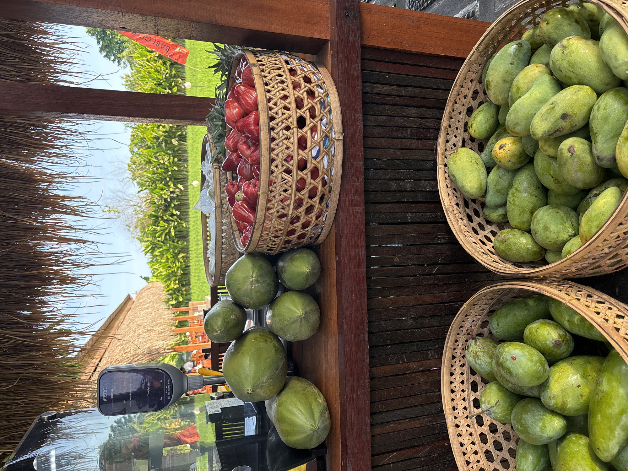 This image depicts a fruit stand in Gianyar, Indonesia, likely on the outskirts of the city given the presence of lush green fields and what appears to be a rice paddy in the background. The fruit stand itself is situated under a thatched roof, supported by wooden pillars.
In the foreground, two woven baskets overflow with green mangoes. To the left of the mangoes, on a wooden surface, sit several large, green papayas. Behind the papayas and to the right, a large woven basket is filled with bright red, bell-shaped fruits, possibly rose apples. A pineapple is visible peeking out from behind this basket.
A digital weighing scale is positioned on the left side of the stand, displaying text in what appears to be Indonesian. The background reveals a vibrant green landscape with rows of plants, suggesting agricultural land. A small red flag with text is planted in the distance, adding a touch of color to the scene. The overall impression is one of a fresh produce market in a tropical setting.