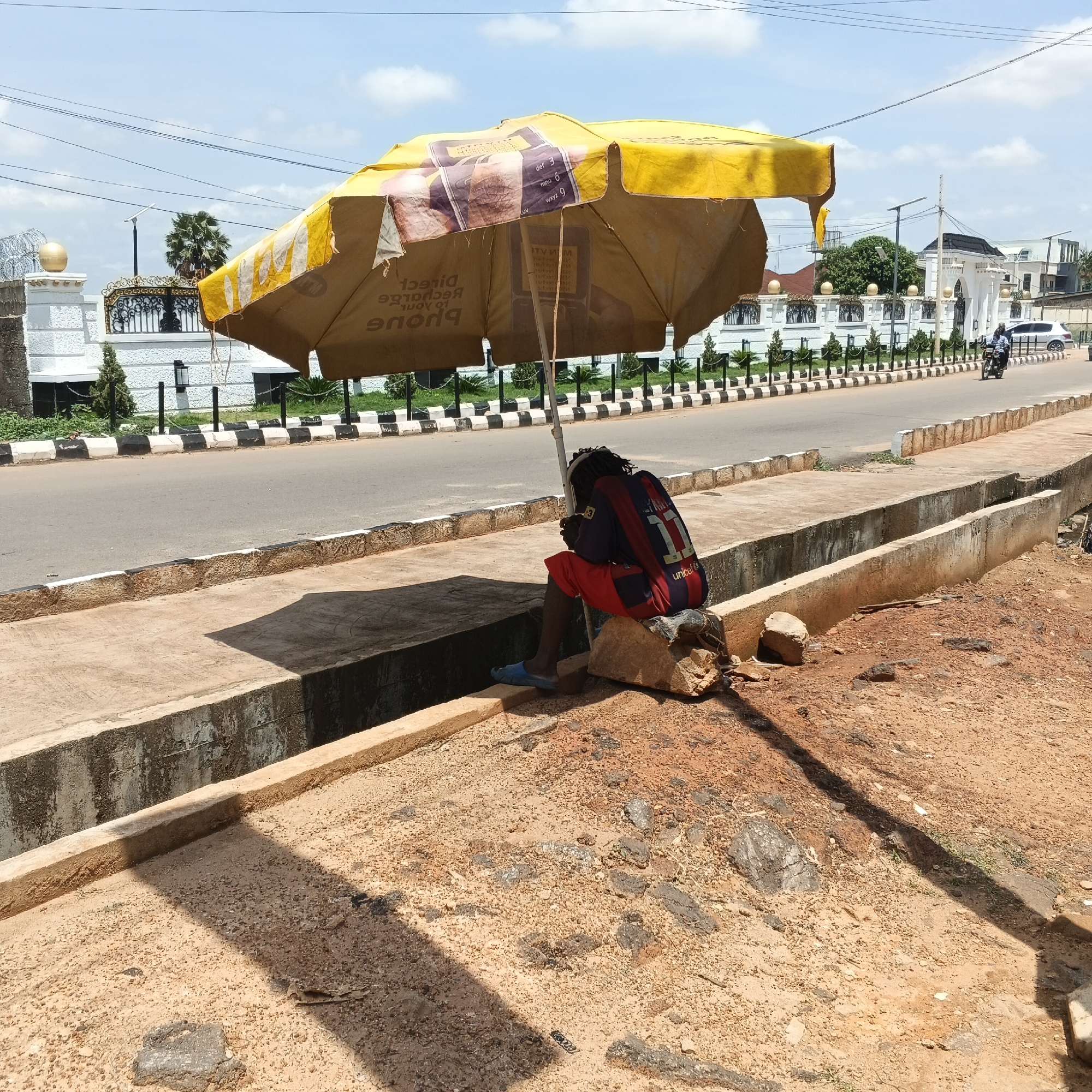 In Shaba, Nigeria, under a faded yellow umbrella, a young individual sits on a rock by the side of a paved road. The individual wears a red t-shirt with the number "11" and blue flip-flops. A backpack is slung over their shoulders. The road is lined with a black and white striped curb and a concrete drainage ditch. In the background, a white building with an ornate gate and a motorbike on the road can be seen. The sky is bright blue with scattered clouds. The ground is covered in sandy soil and small rocks.