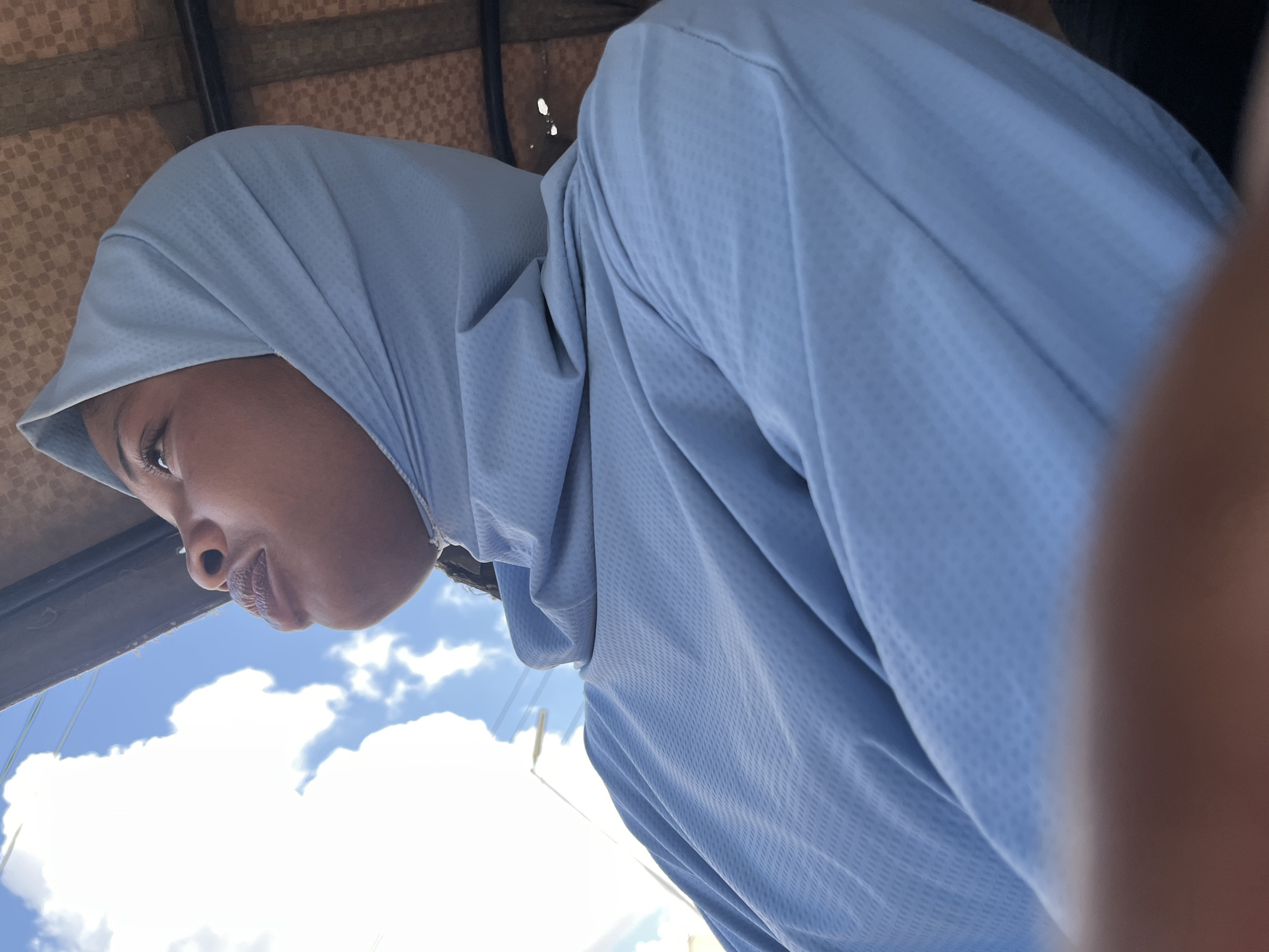 The image captures a close-up, side profile of a young Black individual wearing a light blue hijab and matching long-sleeved shirt. The individual's head is tilted upwards, looking towards a bright sky filled with white clouds. The background suggests an outdoor setting, possibly within a covered area, as indicated by a textured canopy above and to the right of the individual. The lighting suggests a daytime scene. The specific location in Jos, Nigeria, cannot be determined from the visual information provided in the image.