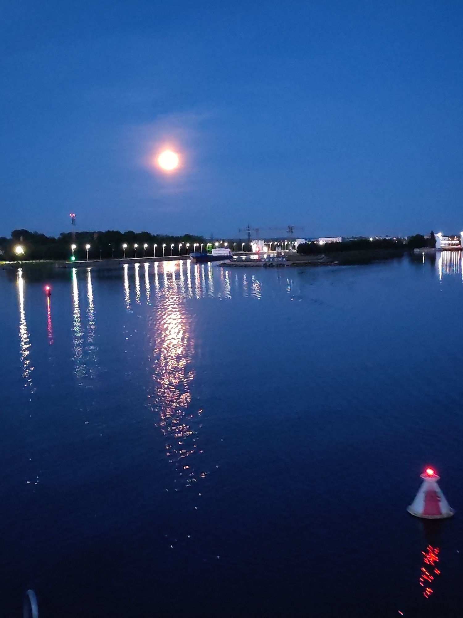 The image shows a body of water at dusk, likely a river or lake, with a dark blue sky above. A bright, full moon is visible in the upper left quadrant of the sky, casting a soft glow. The water reflects the lights from a line of streetlamps and buildings on the far shore, as well as a red and white buoy with a red light in the foreground. The far shore is lined with trees and some industrial or urban structures, including cranes and buildings. The overall atmosphere is serene and peaceful. The location is Sheksna, Russia, along the Sheksna River.