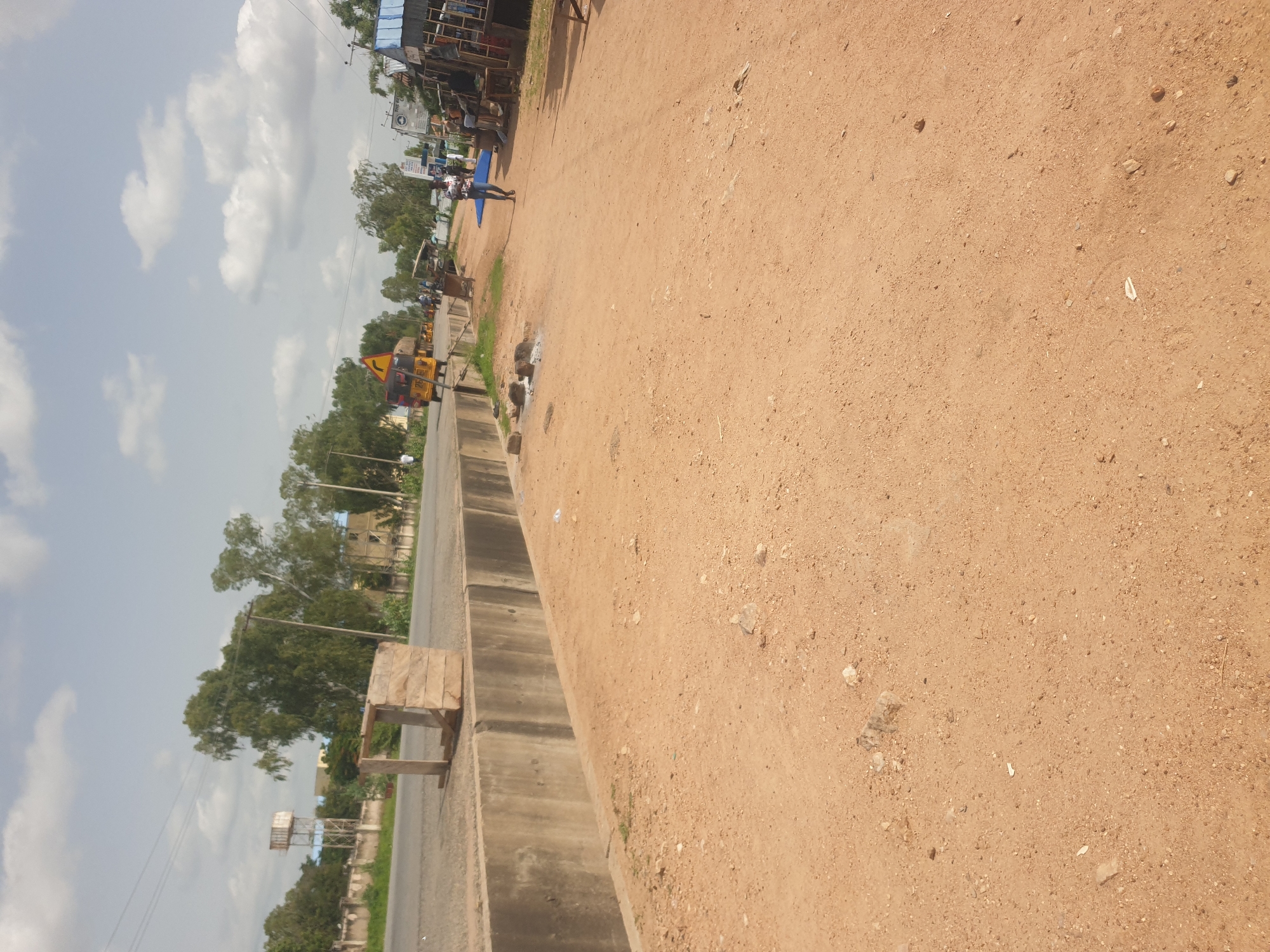 The image depicts a street scene in an outdoor environment under a cloudy sky. A paved road runs horizontally across the frame, bordered by a concrete drainage ditch on the left. On the right side of the road, a sandy area with scattered debris and rocks is visible. Several people are present in the scene. On the road, a yellow autorickshaw is driving away from the camera, and a few other individuals are walking or standing. A person is walking towards the camera on the sandy area, and a group of people are gathered near some market stalls. There are also several market stalls set up along the side of the road, with some appearing to be selling goods. Trees and buildings are visible in the background, suggesting an urban or semi-urban setting. A water tower can be seen in the distance. The overall impression is of a busy street with ongoing commerce and activity.