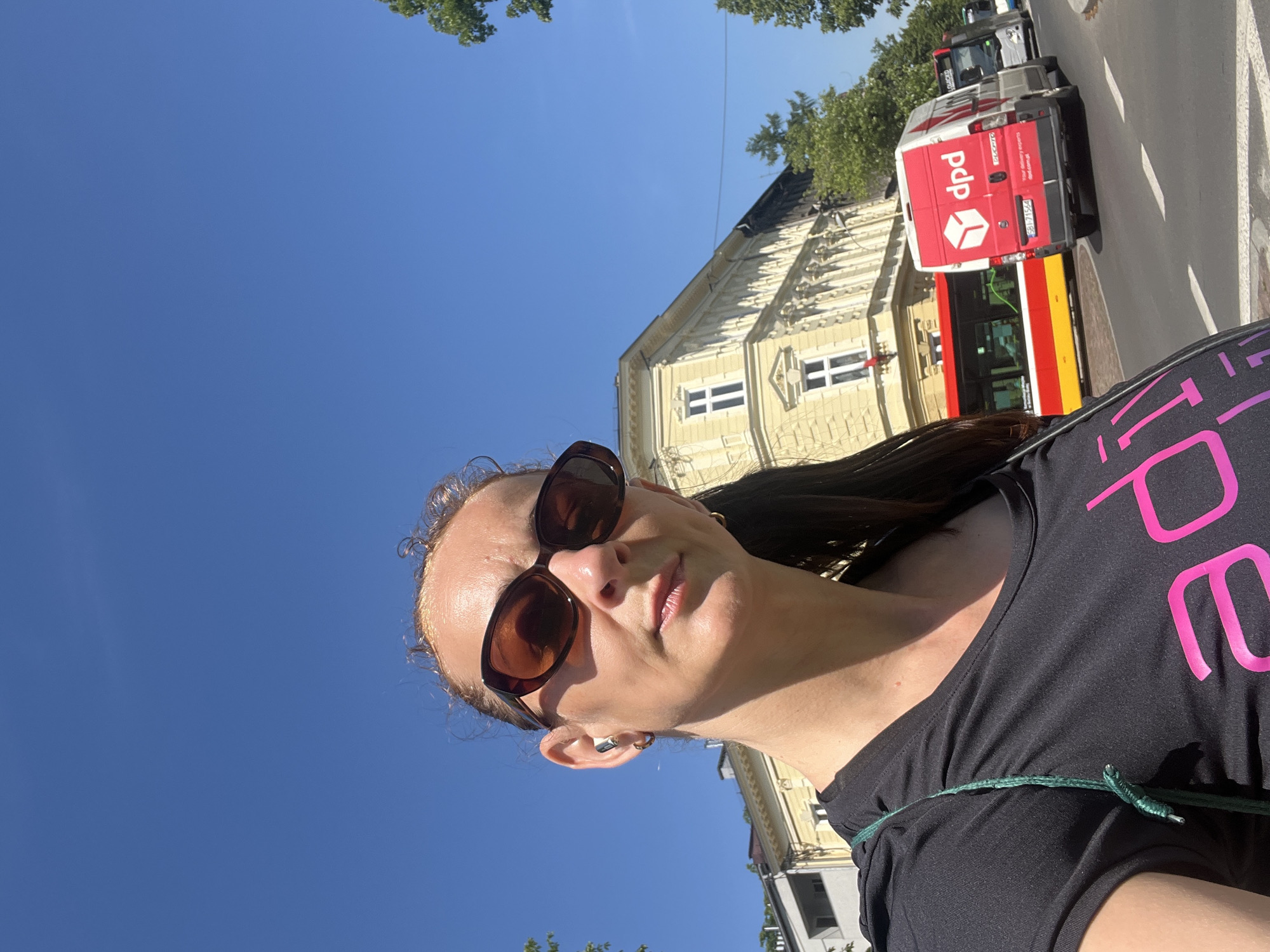 A woman with brown hair pulled back in a ponytail, wearing sunglasses and a black t-shirt with a pink logo, takes a selfie outdoors on a sunny day. She has a green strap from a bag across her shoulder. In the background, a yellow building with white-framed windows is visible, along with trees, a red and yellow bus, and a white DPD delivery van. The scene appears to be a street in a city.