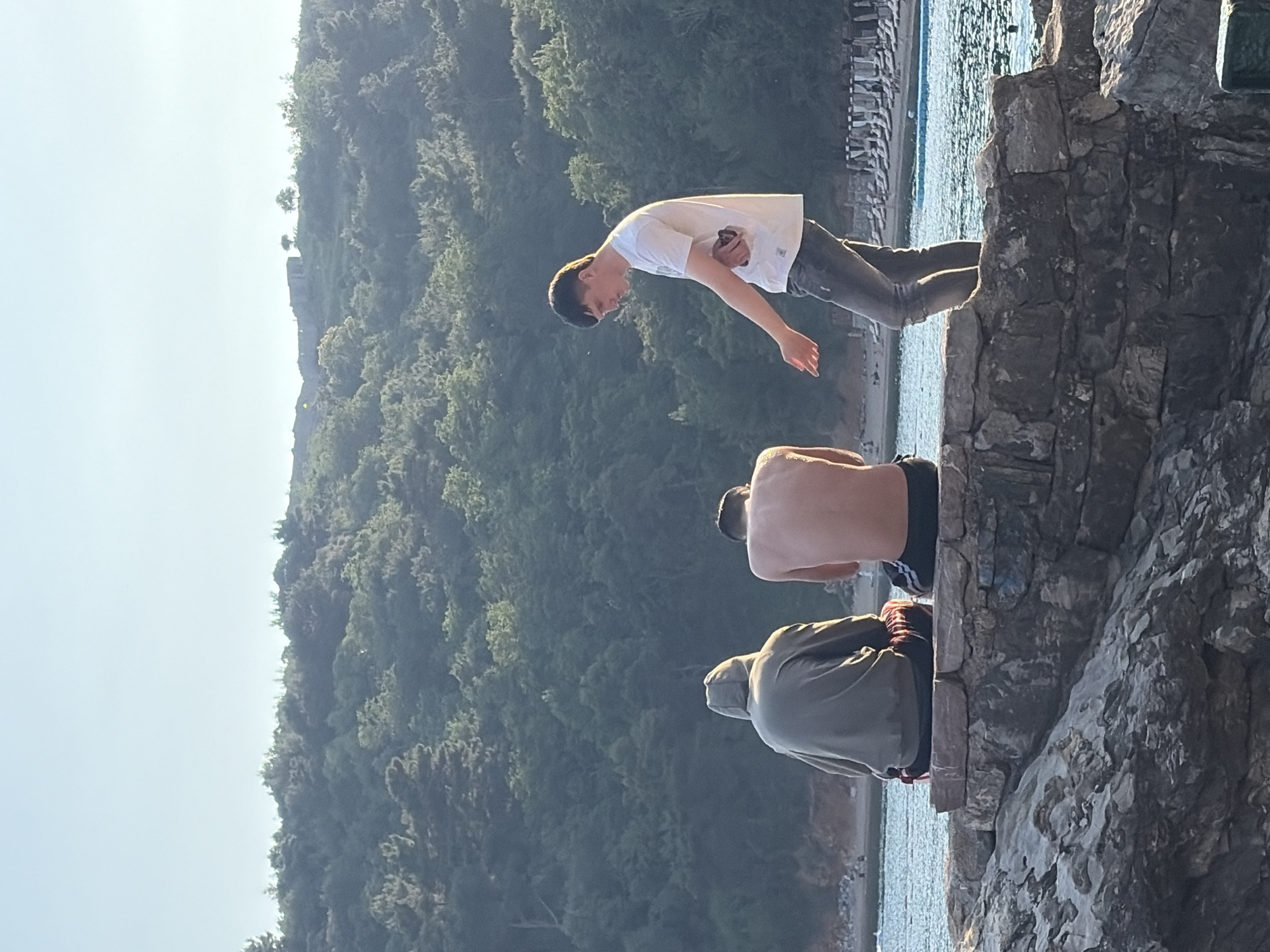Three people are at the edge of a stone structure. One person is leaping into the air toward the water. One person is shirtless. One person is wearing a hoodie. In the background, there is a wooded area with trees and some building structures. The city of Budva, Montenegro is not discernible in this image.