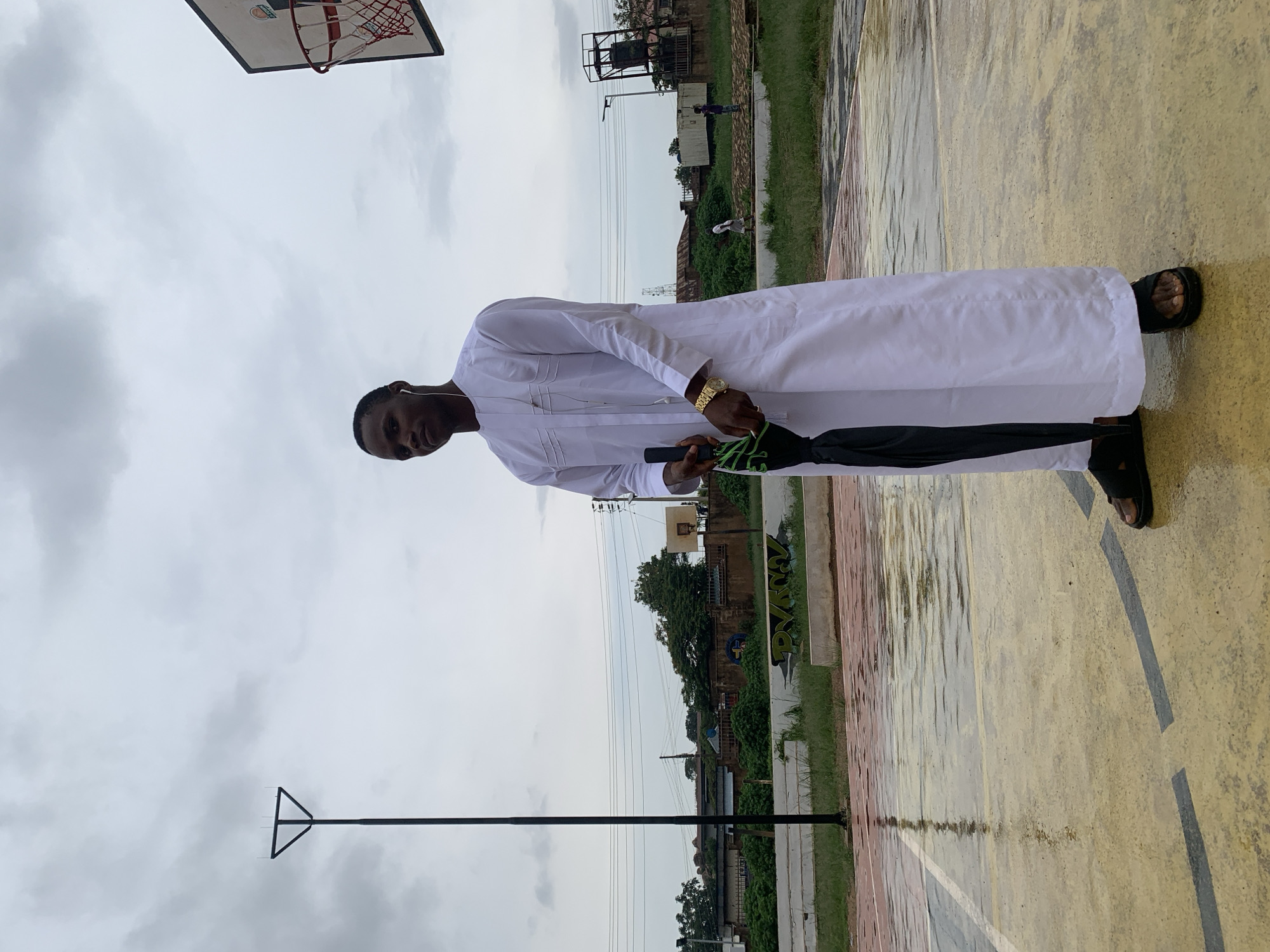 A young man stands on a wet basketball court, wearing a white traditional outfit and black sandals. He holds a closed black umbrella in his left hand and a gold watch is visible on his right wrist. The court has yellow and red markings, and is covered in puddles from recent rain. In the background, there is a basketball hoop, a tall pole with a triangular top, trees, buildings, and power lines. The sky is overcast with grey clouds. The scene appears to be located in Gura Top, Nigeria.
