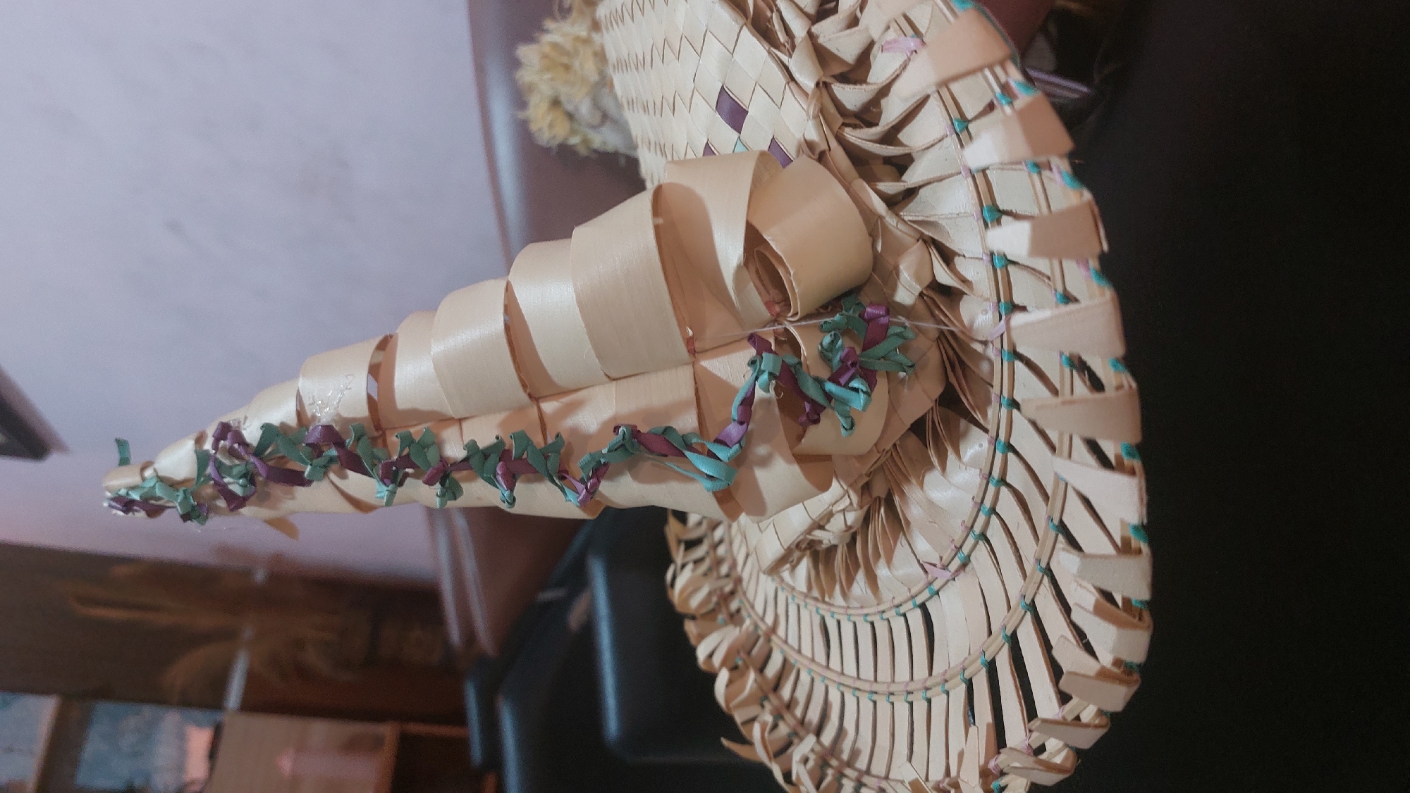 The image shows a close-up of a handmade hat crafted from natural materials, likely palm leaves, characteristic of traditional Indonesian craftsmanship often found in markets or homes in the City of Medan, Indonesia. The hat is conical in shape with a wide brim, meticulously woven. It is decorated with a spiraling ribbon of alternating purple and teal colors, tied in small knots or bows. The brim of the hat features a decorative fringe of pointed, leaf-like elements, also woven from the same natural material, interspersed with small teal beads. The hat is resting on a dark surface, possibly a piece of furniture, with a blurred background that suggests an indoor setting, possibly a living room with furniture and wooden shelves visible. The lighting is dim, and the focus is on the texture and intricate details of the hat.