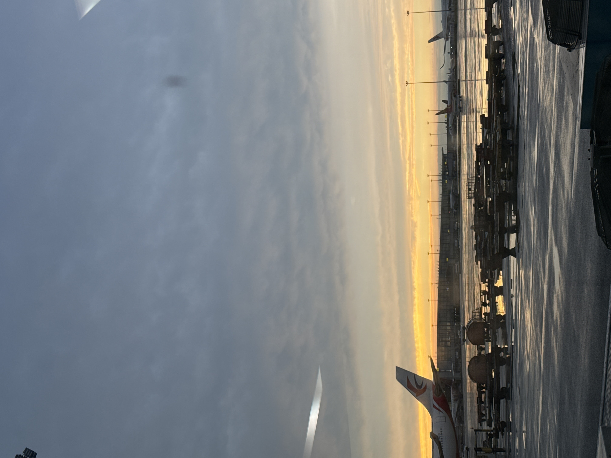 The image depicts a scene at Pudong International Airport in Shanghai, China, during sunrise. The sky is a blend of soft grey clouds and vibrant orange and yellow hues, indicating the early morning light. Several commercial airplanes are visible on the tarmac, with one prominent airplane in the foreground featuring a white fuselage with red and orange accents on its tail fin. The ground area is wet, reflecting the light and suggesting recent rain or dew. Various airport equipment, including baggage carts and service vehicles, are scattered across the tarmac. Tall lampposts with multiple lights are positioned throughout the airport grounds. In the distance, airport terminals and other aircraft can be observed. The overall atmosphere is calm and quiet, typical of an airport at dawn.