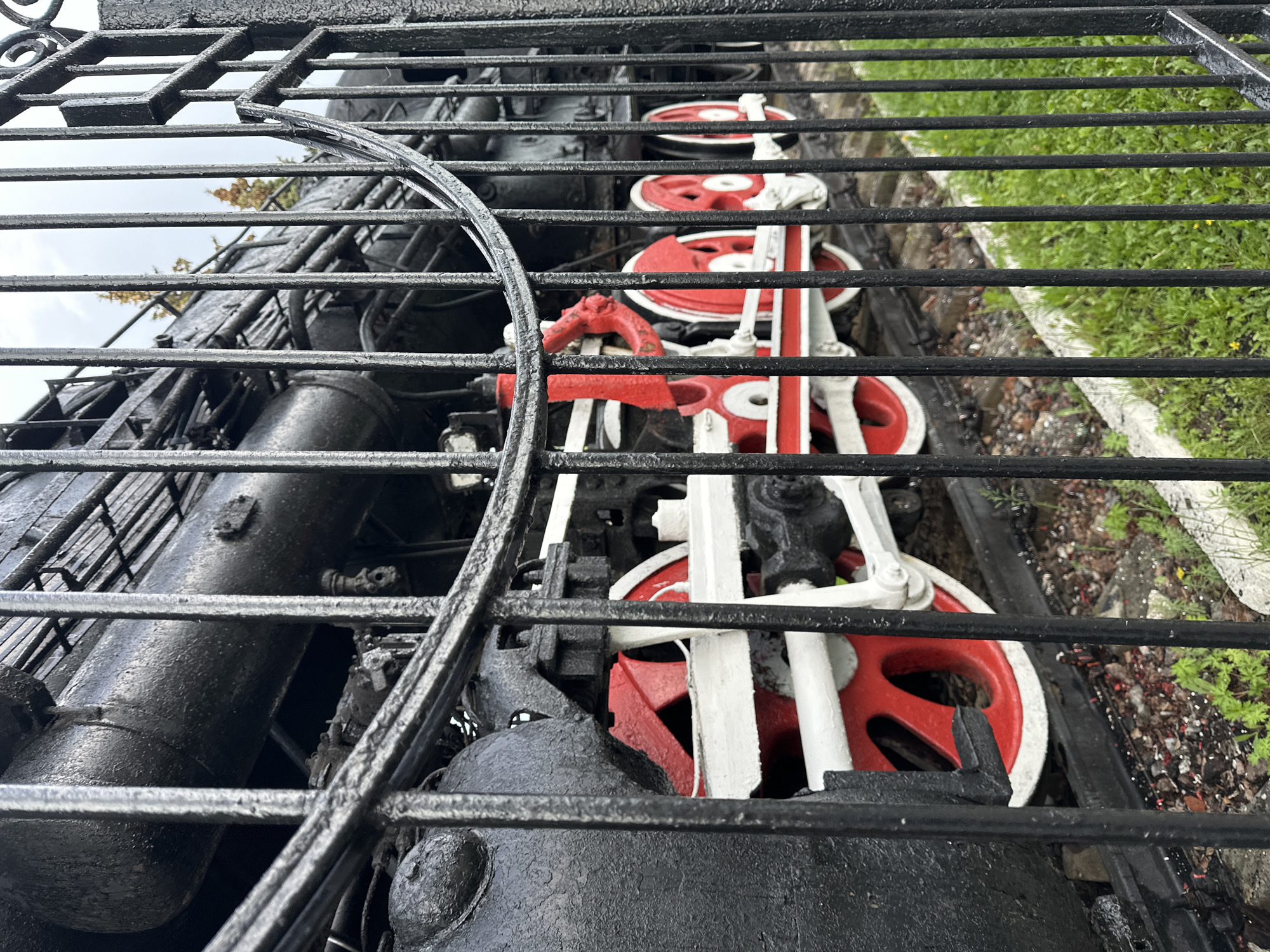 The image captures a close-up view of a vintage steam locomotive's wheels and machinery, partially obscured by a black metal fence or railing. The locomotive is painted in a color scheme of black, red, and white. The red and white are prominently featured on the wheels and connecting rods. The black parts appear to be the engine and other mechanical components. The machinery is set against a backdrop of greenery and a cloudy sky. The fence appears weathered, possibly due to age and exposure to the elements. The location is likely Vladimir, Russia, based on the visual elements.