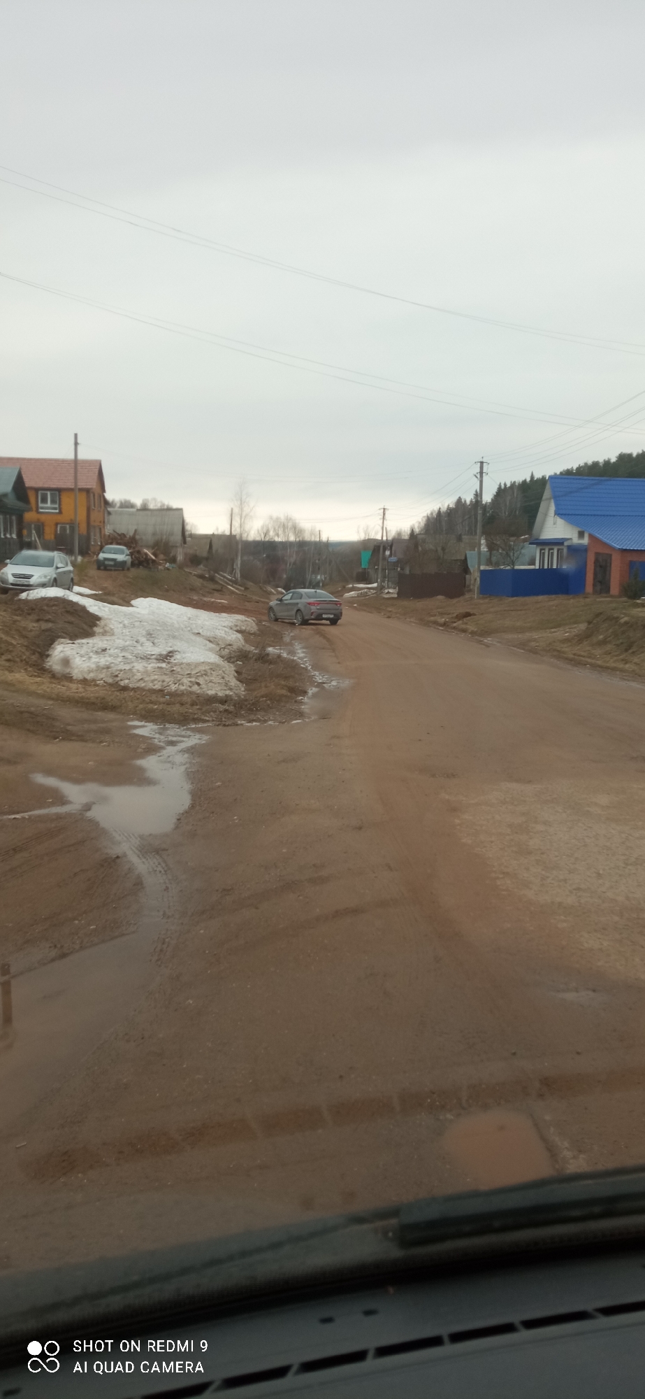 The image depicts a dirt road in what appears to be a rural area of Ольховка, Russia. The sky is overcast. Patches of melting snow are visible on the roadside. Several houses are situated along the road, some with colorful roofs (blue and orange). At least three cars are visible: one gray sedan on the road and two more parked. Power lines run overhead. The image is taken from inside a vehicle, with part of the dashboard visible.