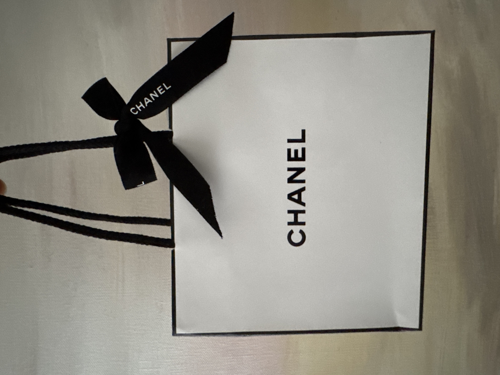 A white Chanel shopping bag with black handles is held against a textured light-colored background. A black Chanel ribbon is tied in a bow on the right side of the bag's handles, with the word "CHANEL" printed in white on the ribbon. The word "CHANEL" is also printed in large black letters in the center of the shopping bag. The image appears to be an indoor shot, with no discernible city or location details.