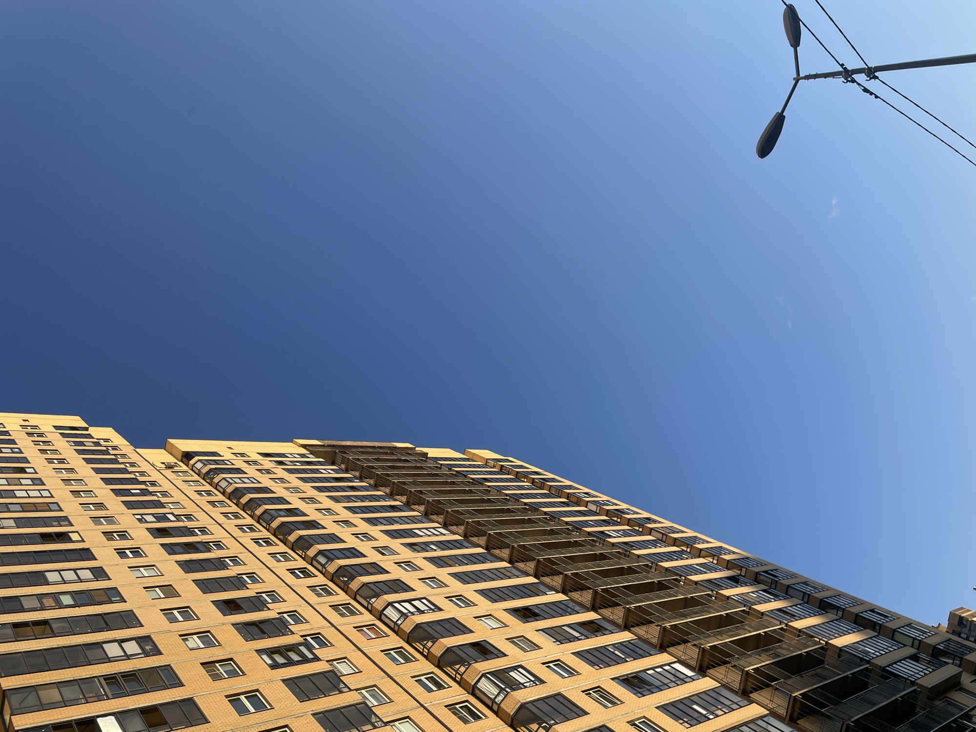 An upward, angled shot captures a modern, beige apartment building with numerous windows and balconies on the left side of the frame. The building is bathed in the warm, golden light of the sun, casting long shadows. The right side of the frame is dominated by a clear, bright blue sky, with only a faint wisp of a cloud visible. In the lower right corner, a street lamp with two lights stands, connected by wires to poles that extend out of the frame. The scene is a typical urban landscape. This image was taken in Pargolovo, Russia.