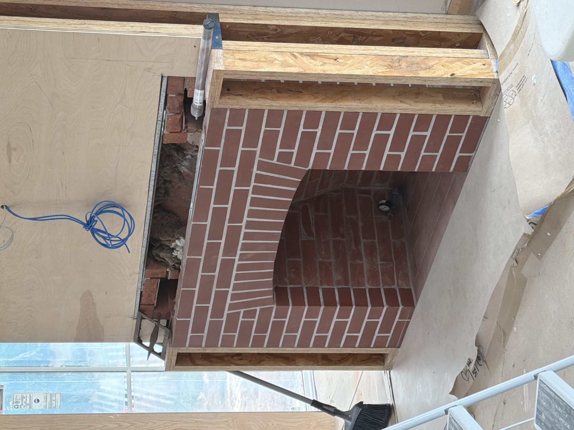 This media file captures a construction scene focused on a partially built brick fireplace in what appears to be a residential interior. The fireplace features a distinct arched opening and is constructed with red bricks laid in a stretcher bond pattern with white mortar. Above the brickwork, exposed wooden framing, likely OSB (Oriented Strand Board) and lumber, is visible, indicating the ongoing construction of the surrounding structure. A rolled-up bundle of blue electrical wire is loosely coiled on a plywood surface to the left of the fireplace. To the right, a large sheet of brown paper, possibly for protection, is draped over a section of the construction, with some text and graphics visible on its surface. A broom with a long handle is leaning against a window in the lower part of the frame, suggesting that cleaning or site preparation is underway. The lighting suggests it is daytime, likely indoors, with natural light coming from a window. The scene is devoid of people but implies human activity through the presence of construction materials and tools. The overall impression is one of a home renovation or new build project in progress.