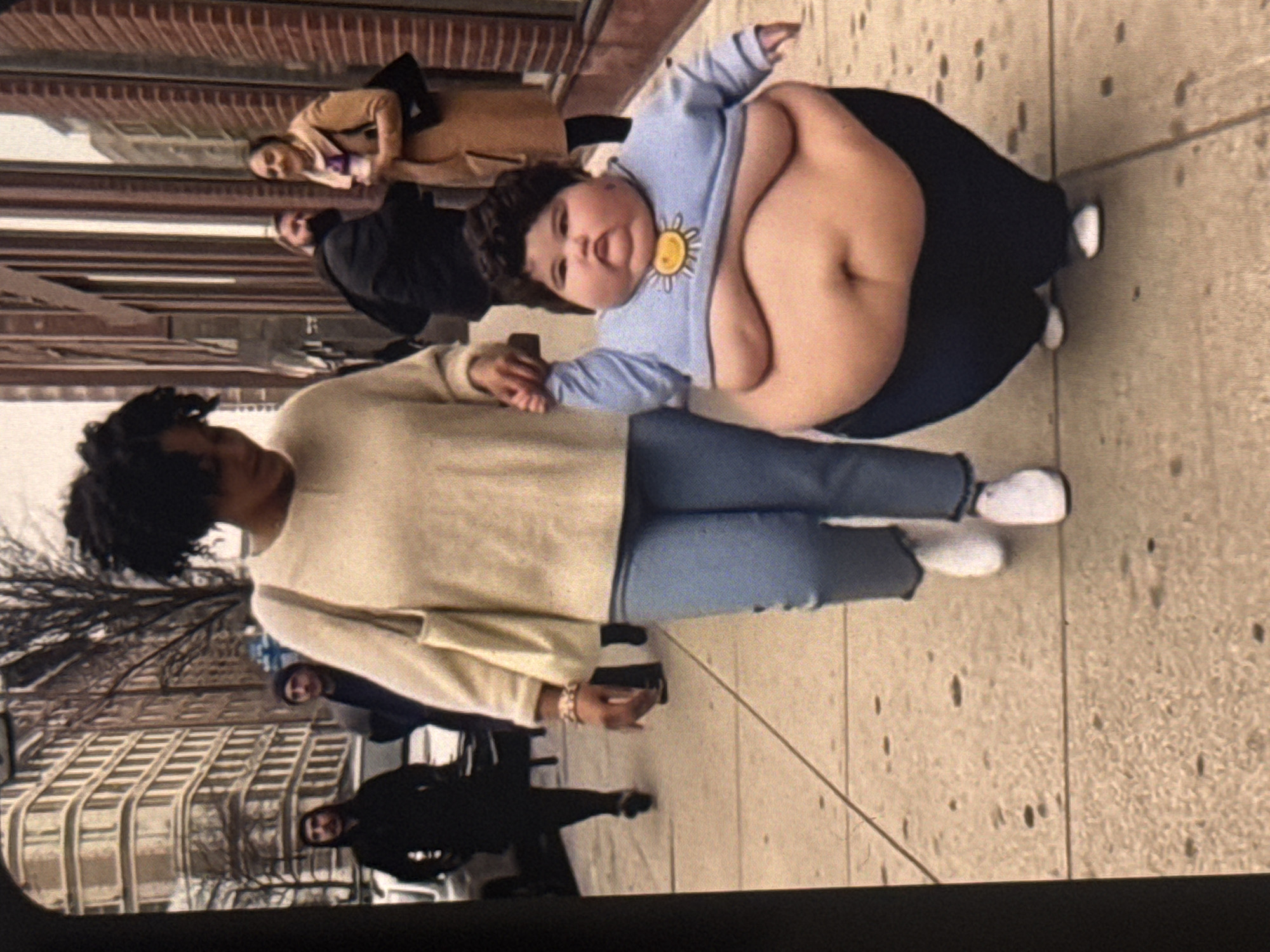 A woman with dark curly hair, wearing a cream-colored sweater and blue jeans, walks hand-in-hand with a very overweight child on a sidewalk. The child is wearing a light blue long-sleeved shirt with a yellow sun emblem and dark pants. In the background, other people are walking, and buildings line the street. The scene appears to be a city street. The location is not Kuta Selatan, Indonesia.