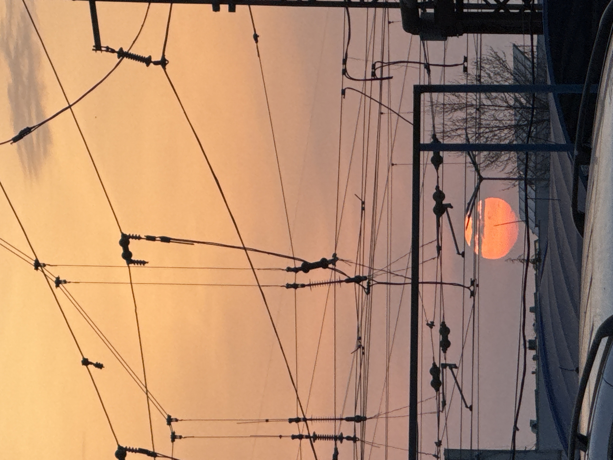 The image depicts a sunset in Burgas, Bulgaria, obscured by power lines and infrastructure related to electric transportation, potentially trams or trains. The setting sun appears as a blurry orange disc, partially hidden behind the lines. A steel framework structure, likely part of the electric transport system, is visible. The background sky exhibits a gradient of orange and light pink hues indicative of sunset. Parts of parked cars are visible on the right side of the image, indicating an urban environment. Bare branches of a tree are also visible in the background.