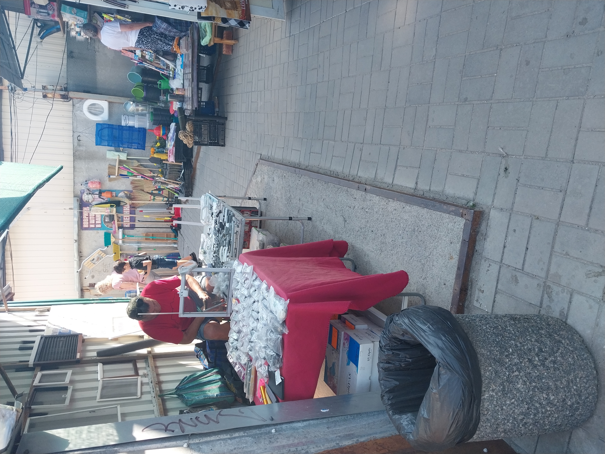 In a street market setting in Dnipro, Ukraine, a man in a red t-shirt and shorts is seated at a table covered with a red tablecloth. He is examining small items laid out on the table, which are contained within clear plastic bags. Numerous such bags are spread across the tablecloth, suggesting they contain small goods for sale or display. Beside the table, a concrete trash receptacle lined with a black plastic bag is visible. In the background, a woman and a child are standing, with various market stalls and merchandise, including cleaning supplies and household items, visible further down the street. The ground is paved with interlocking grey bricks.