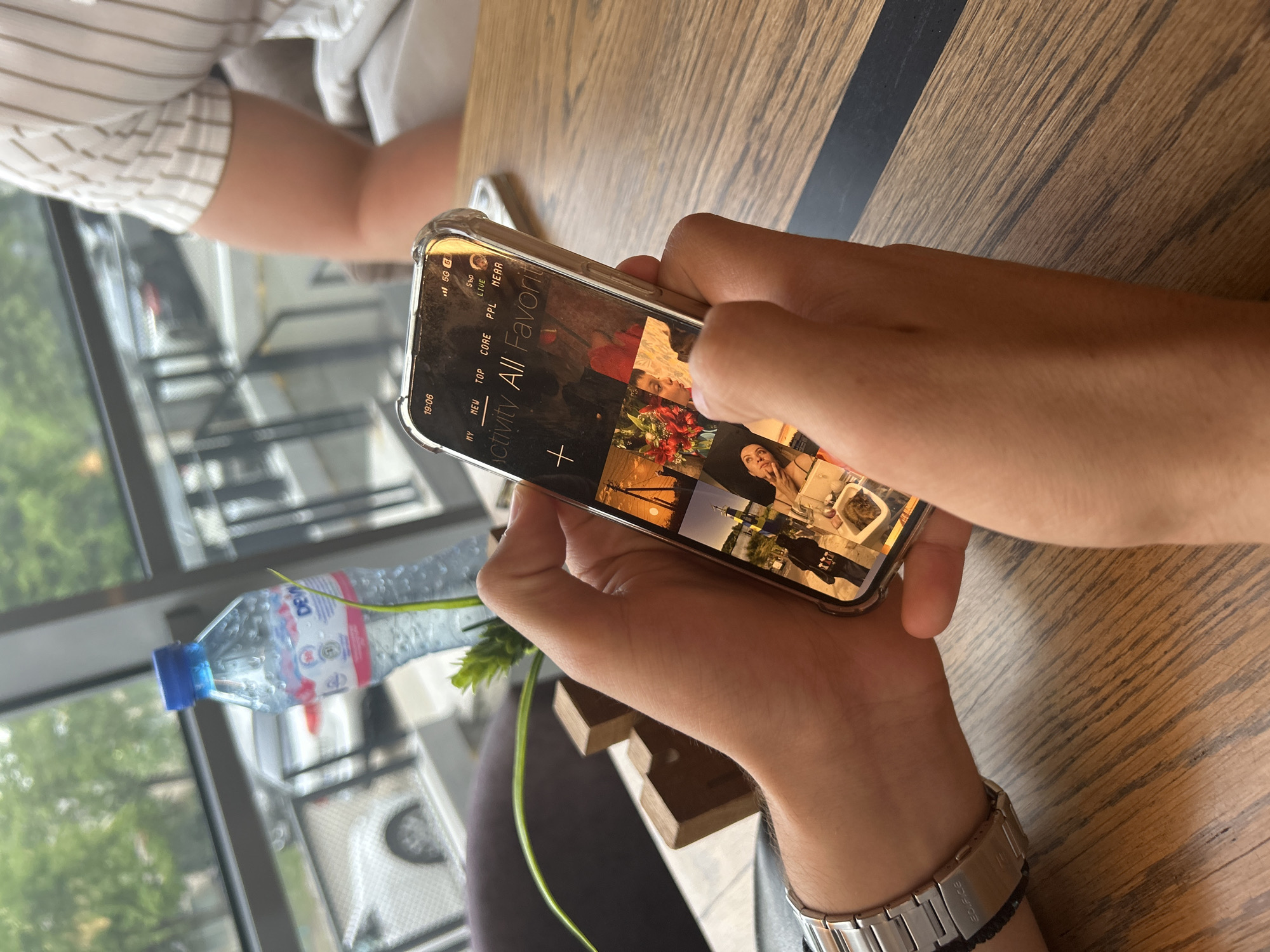 The image, captured from a skewed, close-up perspective, centers on a person holding a smartphone with both hands, positioned over a wooden table with a black stripe. The phone, encased in a clear cover, displays a grid of thumbnail images, suggesting a photo gallery or a social media feed. The interface includes categories such as "MY", "NEW", "TOP", "CORE", "PPL", "NEAR", and a prominent title "Activity All Favorit", along with a "+" icon. The screen also shows the time "19:06", network indicators "5G G4", and text including "ГъP" and "LIVE".
The person's forearms and hands are visible, with a silver-colored watch, bearing the brand "EDIFICE" on its clasp, worn on the left wrist. Resting on the table beside the phone is a clear plastic water bottle with a blue cap, featuring the brand "DEW" on its label, and a green plant stem.
In the blurred background, another person wearing a striped shirt is partially visible, seated at what appears to be the same table. Behind them, a large window reveals an outdoor scene with green foliage, indicating daylight outside. The setting suggests an indoor establishment, possibly a cafe or restaurant, in Sofia, Bulgaria, during the early evening hours.