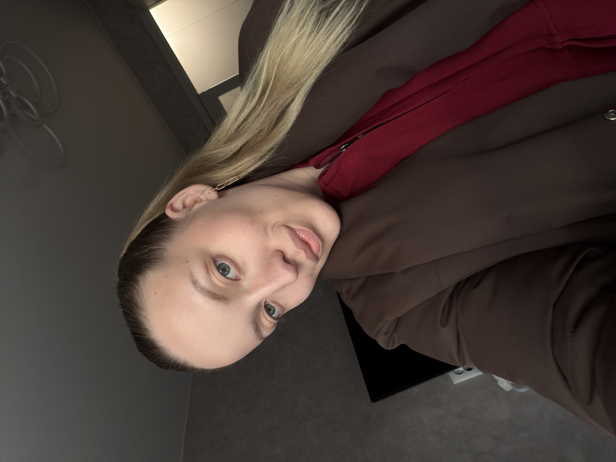 A young woman with fair skin and blonde hair is looking at the camera. She is wearing a brown jacket and a red top. The background appears to be an interior space, with a grey wall. The image appears to be taken indoors with an overhead light source visible. No evidence suggests the location of Reshetnikovo, Russia.