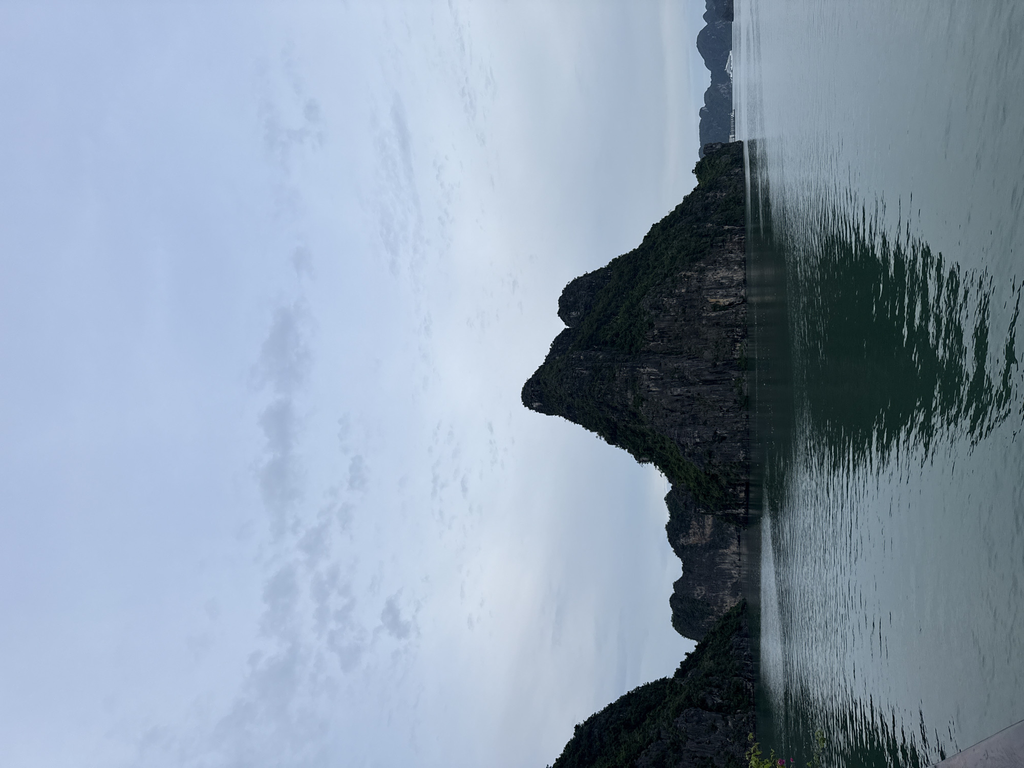 A wide shot captures the limestone karsts of Ha Long Bay, Vietnam, a UNESCO World Heritage site located within the administrative boundaries of the city of Hai Phong. The karsts, covered in lush green vegetation, rise dramatically from the calm, teal-colored waters of the bay. The sky above is overcast with a pale blue hue, with wispy clouds scattered across it, suggesting either early morning or late afternoon. In the distance, a few tour boats are visible, adding a sense of scale and hinting at the bay's popularity as a tourist destination. The water reflects the muted light and the silhouettes of the karsts, creating a serene and somewhat dramatic scene. The overall impression is one of natural beauty and geological grandeur.