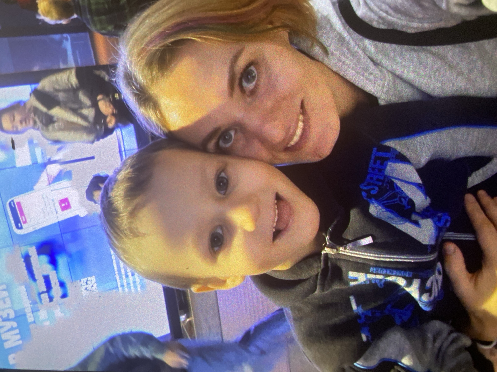 A candid, warm portrait features a woman and a young child, likely a mother and son, captured indoors at what appears to be a museum in Saint Petersburg, Russia. The woman, with blonde hair and highlights, smiles genuinely at the camera, her blue eyes bright. She wears a grey hooded sweatshirt. The child, a young boy with light hair, rests his head close to hers, looking at the camera with a wide, joyful smile and his tongue slightly out, conveying playfulness. He is wearing a dark grey or black hooded sweatshirt with blue and white graphic text that partially reads "STREET" on the front.
The background is softly blurred but reveals large digital display screens with glowing blue light. On one screen in the lower left, the Russian word "МУЗЕЙ" (Museum) is clearly visible, providing a strong location cue. Another screen shows what looks like a mobile application interface or a diagram. Further back, out-of-focus figures of other people can be seen, suggesting a public environment. The lighting is artificial, consistent with an indoor setting like an exhibition or a modern museum. The overall mood of the image is happy and affectionate.