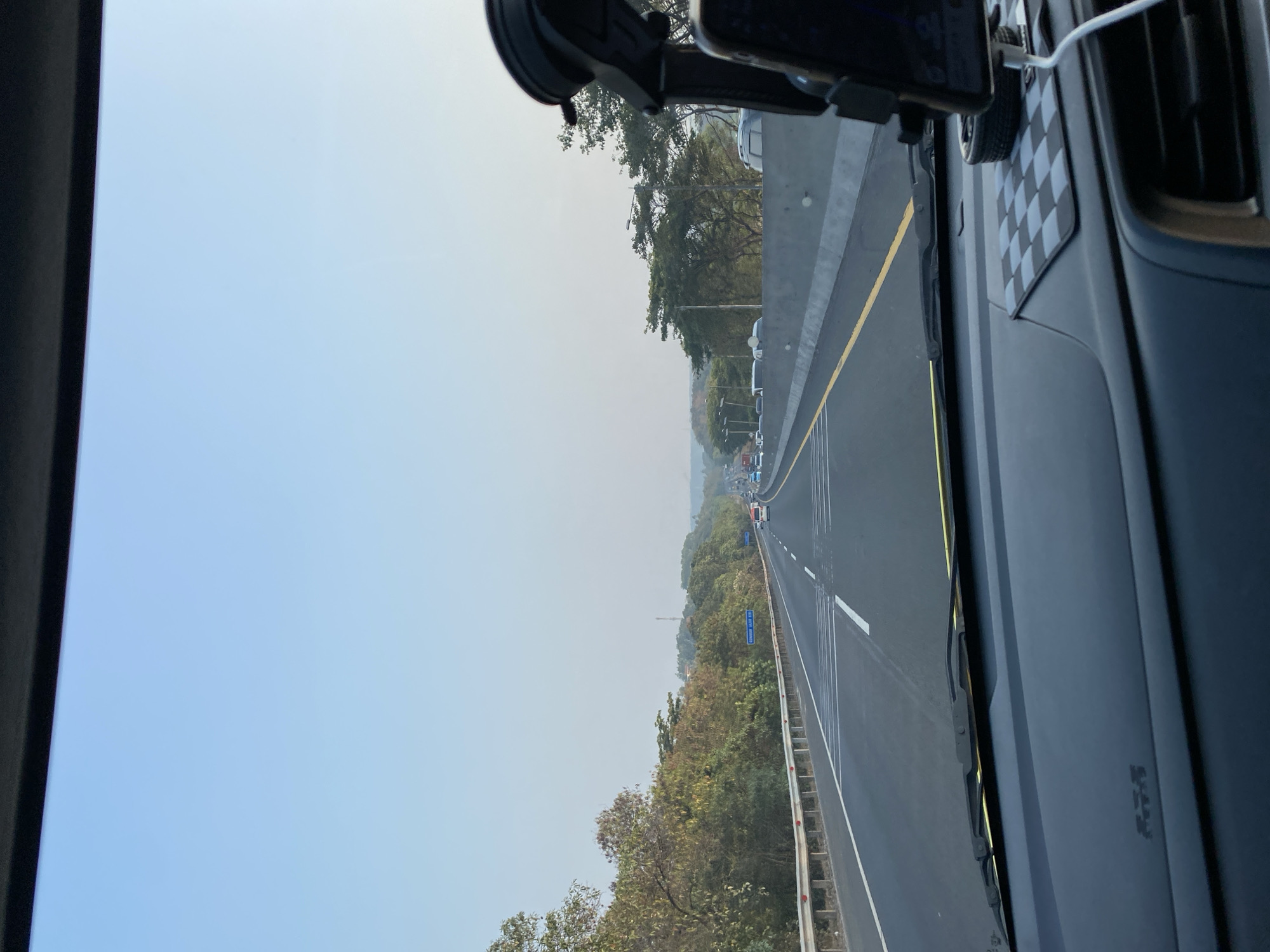 The image shows a view from inside a vehicle on a highway. The road is paved with clear lane markings and a yellow line separating the lanes. The highway is flanked by trees and vegetation on the left and a concrete barrier on the right. In the distance, other vehicles are visible on the highway, indicating moderate traffic. The sky is clear and blue, suggesting it is daytime. A cell phone mount is attached to the windshield or dashboard on the right side of the frame. The interior of the car is visible in the foreground, including the dashboard and windshield wipers. This scene is consistent with the Cipularang Toll Road, a major highway connecting Jakarta and Bandung, which passes through the city of Purwakarta, Indonesia.