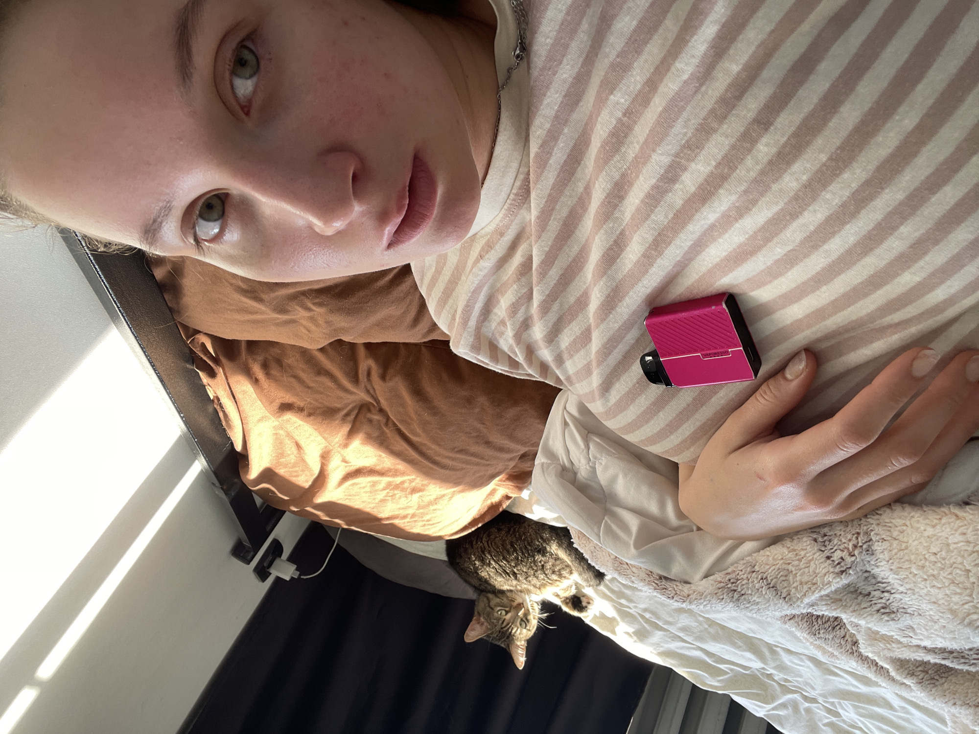 The image depicts a woman lying in bed, partially obscured by blankets. A tabby cat is visible in the background, also on the bed. A pink vape device rests on the woman's chest. The environment appears to be a bedroom, with sunlight casting shadows from blinds onto the wall and ceiling. The scene is consistent with a domestic setting, and no specific elements suggest a location within the city of Chaikovsky, Russia.